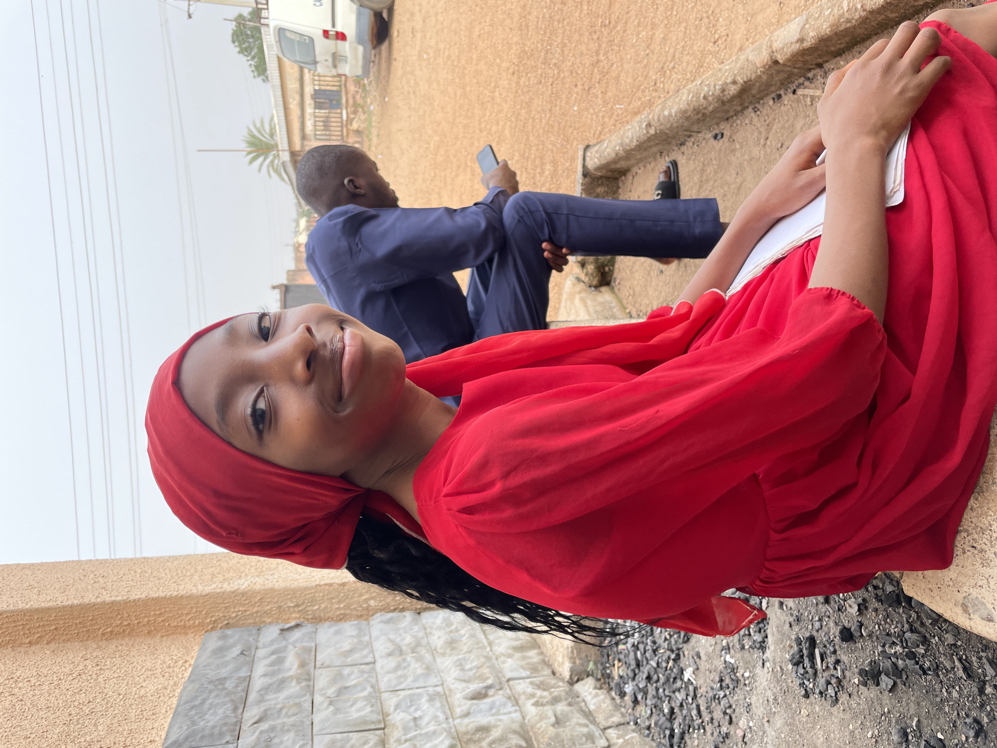 The image shows a woman in a red dress and headscarf sitting on a ledge. She is holding a white object. Behind her, a man in a blue suit sits with a phone in his hand. A white van is visible in the background. The setting appears to be outdoors in Jos, Nigeria, based on the environment and clothing styles.