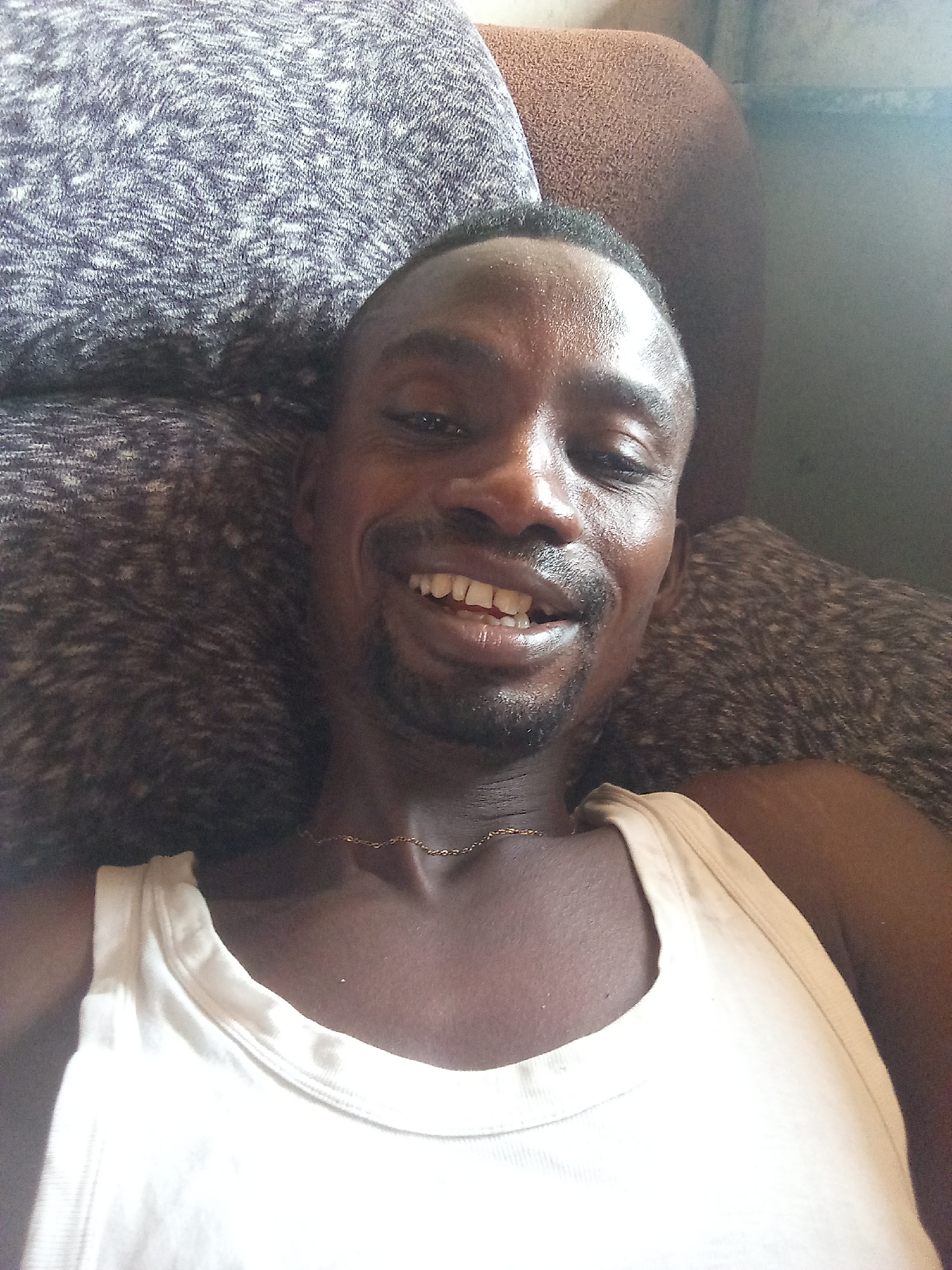 A close-up, upward-angled shot captures a dark-skinned man with short dark hair and a trimmed beard or stubble, smiling broadly directly at the camera. His teeth are visible in his wide, cheerful grin. He is wearing a plain white sleeveless undershirt, and a thin gold chain is visible around his neck, resting on his collarbone.
The man appears to be reclining or lying down on soft furniture, likely a couch or bed. Behind him, textured cushions or pillows in shades of dark gray and brown provide a soft background. A lighter brown, textured surface, possibly another part of the furniture, is visible on the upper right side of the frame. The lighting suggests an indoor setting during the daytime. The man's expression conveys happiness and relaxation. No visible text is present in the image. The scene depicts a casual moment in what appears to be a domestic environment in Adonkolo, Nigeria.