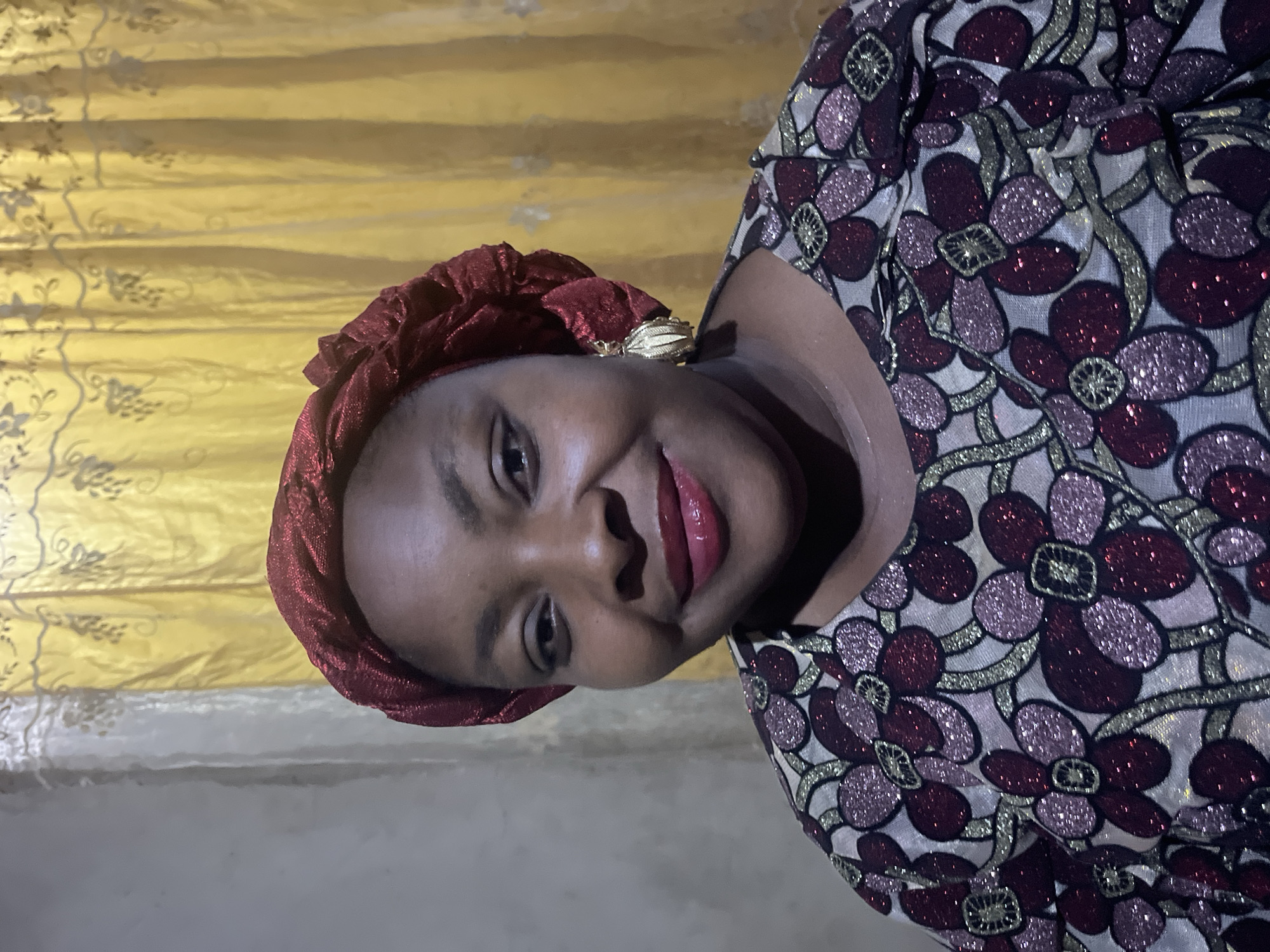 A medium shot portrait shows a woman smiling and looking at the camera. She has dark skin and is wearing a red headscarf and a patterned top with red and pink flowers outlined in gold. She is also wearing a gold earring on her right ear. The background consists of a yellow curtain with a floral pattern and a grey wall. The lighting is dim, suggesting an indoor setting at night or in low light conditions. The woman's makeup is noticeable, with bright red lipstick and what appears to be eyeliner.