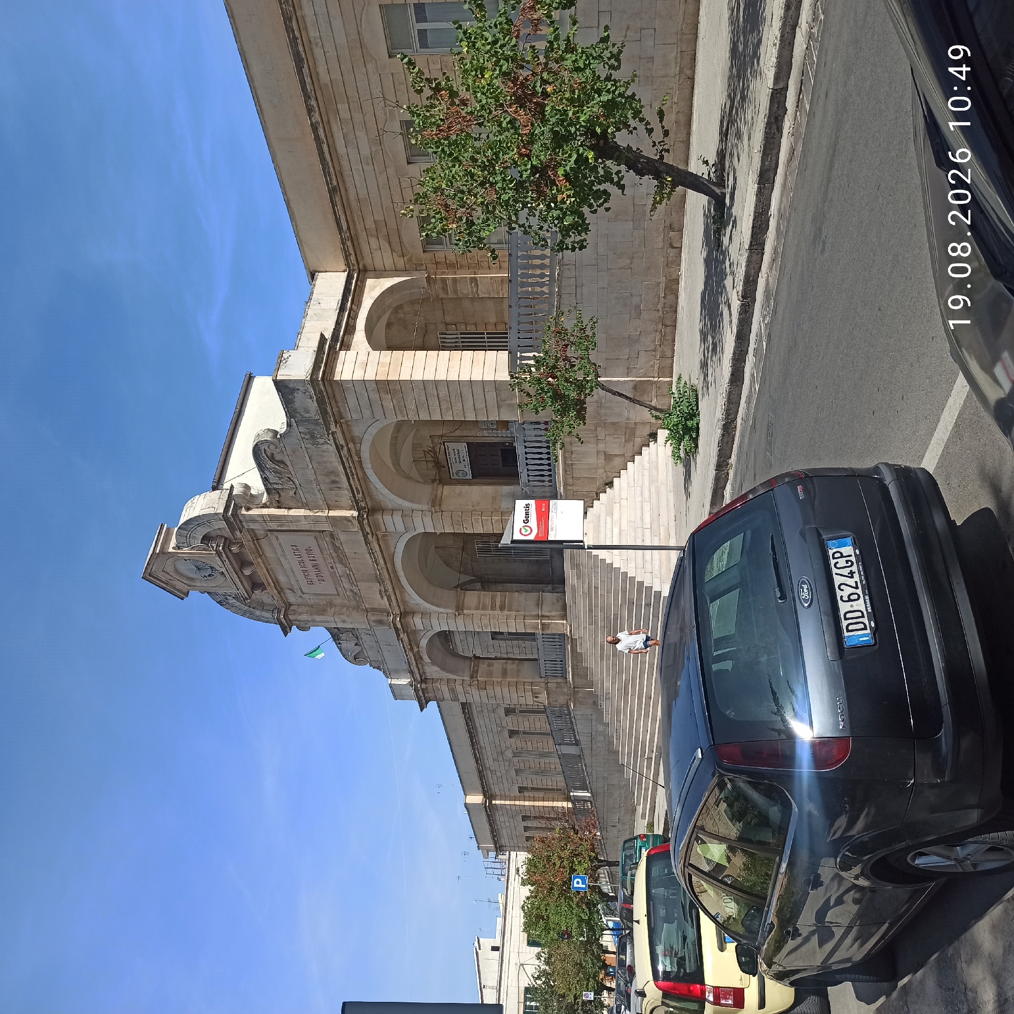 The image shows the exterior of a stone building with arched entrances and a prominent clock tower. The building appears to be an educational institution, as indicated by the sign above the main entrance that reads "EDIFICIO SCOLASTICO GIOVANNI BOVIO." A green and white flag is visible on the upper part of the building. In front of the building, a wide set of stone stairs leads up to the entrance. A person in a white t-shirt and blue shorts is walking down the stairs.
The street in front of the building is lined with parked cars, including a dark grey Ford Focus with license plate "DD 624GP" in the foreground, and a yellow car to its left. Trees with green foliage are planted along the sidewalk. The sky is clear and blue, suggesting a sunny day.
The timestamp in the lower right corner of the image indicates "19.08.2026 10:49." This image is located in Ruvo di Puglia, Italy, a town in the Metropolitan City of Bari, in the Apulia region.