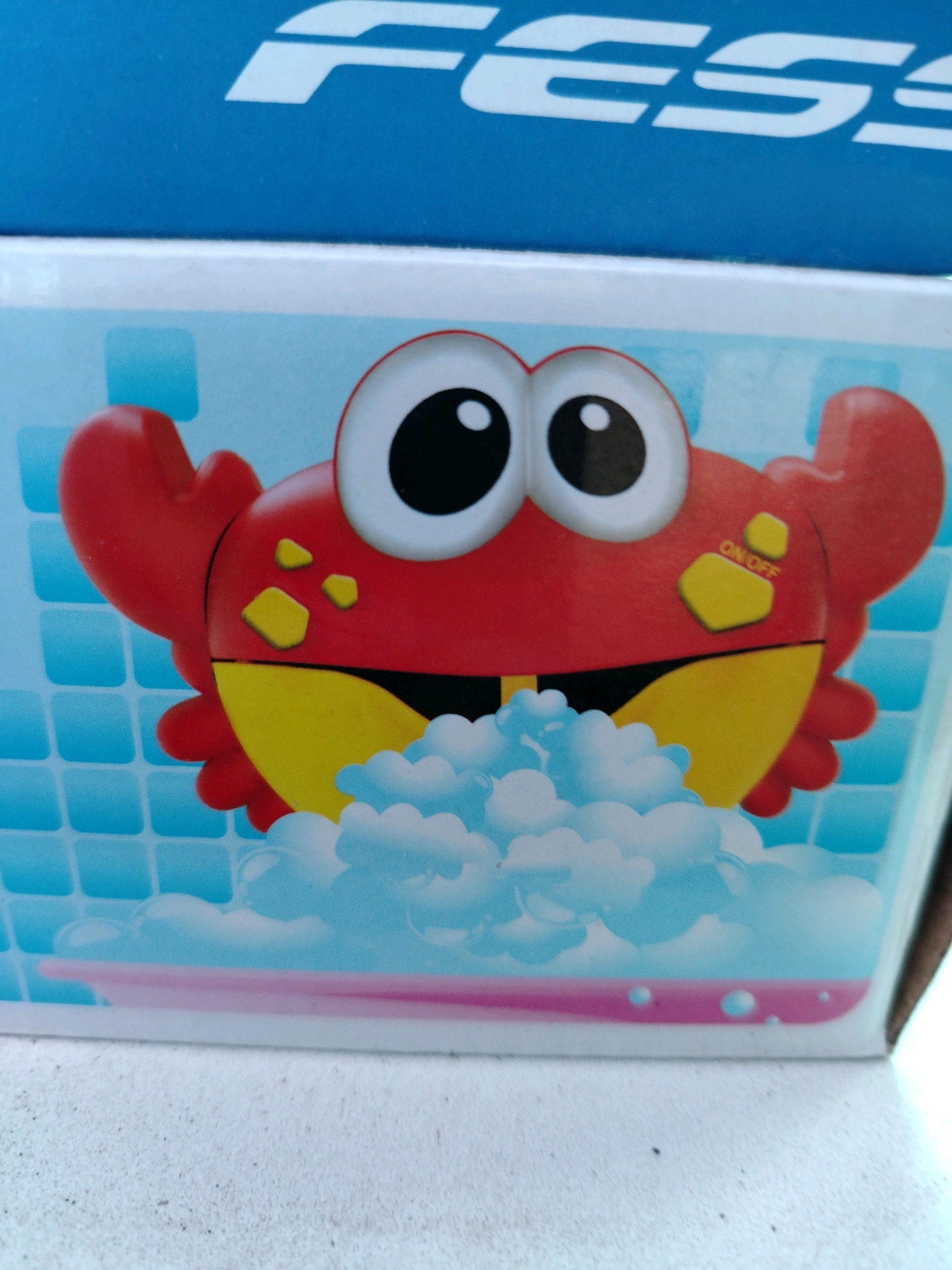 A close-up, eye-level, vertical, outdoor shot shows a portion of a blue and white cardboard box with a cartoon image of a red crab with yellow accents and large googly eyes. The crab is in front of a light blue background with a grid pattern resembling bathroom tiles. White, fluffy foam is depicted around the crab's mouth, suggesting it is a bubble-making toy. A pinkish-purple band runs across the bottom of the box. The box appears to be placed on a textured, off-white surface, possibly a sidewalk or a rough tabletop, with some dark specks visible on it. The background of the image shows the blue portion of the box with white stylized text that reads "FESS" in a partial view. The location is not discernible from the image and the mention of Pregradnoye, Russia is not supported by visual evidence.
