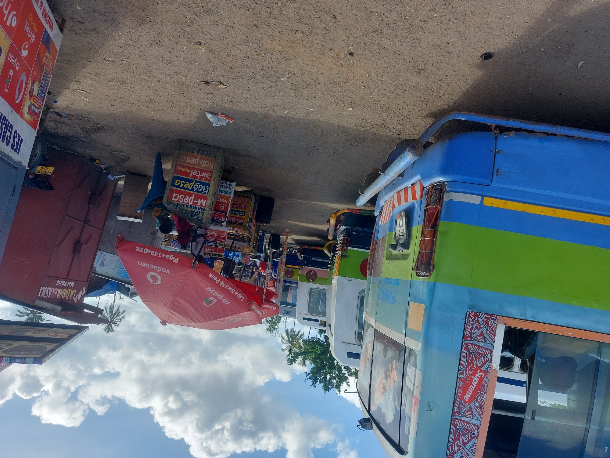 The image depicts a street scene in Dar es-Salaam, Tanzania, characterized by a row of vibrant public transportation vehicles, known locally as "dala dalas". The dala dalas, painted in bright shades of blue, green, and white, are parked along the roadside. Some feature advertising decals, one notably with a "Supreme" logo. An open-air market with vendors selling mobile phone airtime and M-Pesa services is visible. Branded umbrellas and stacked boxes displaying logos of various mobile money operators (Vodacom, Tigo Pesa, Airtel Money, M-Pesa) are present. A red-roofed shop with the sign "Duka la dawa za Asili" is visible. The sky above has a mix of blue and puffy white clouds. Debris is scattered on the ground.