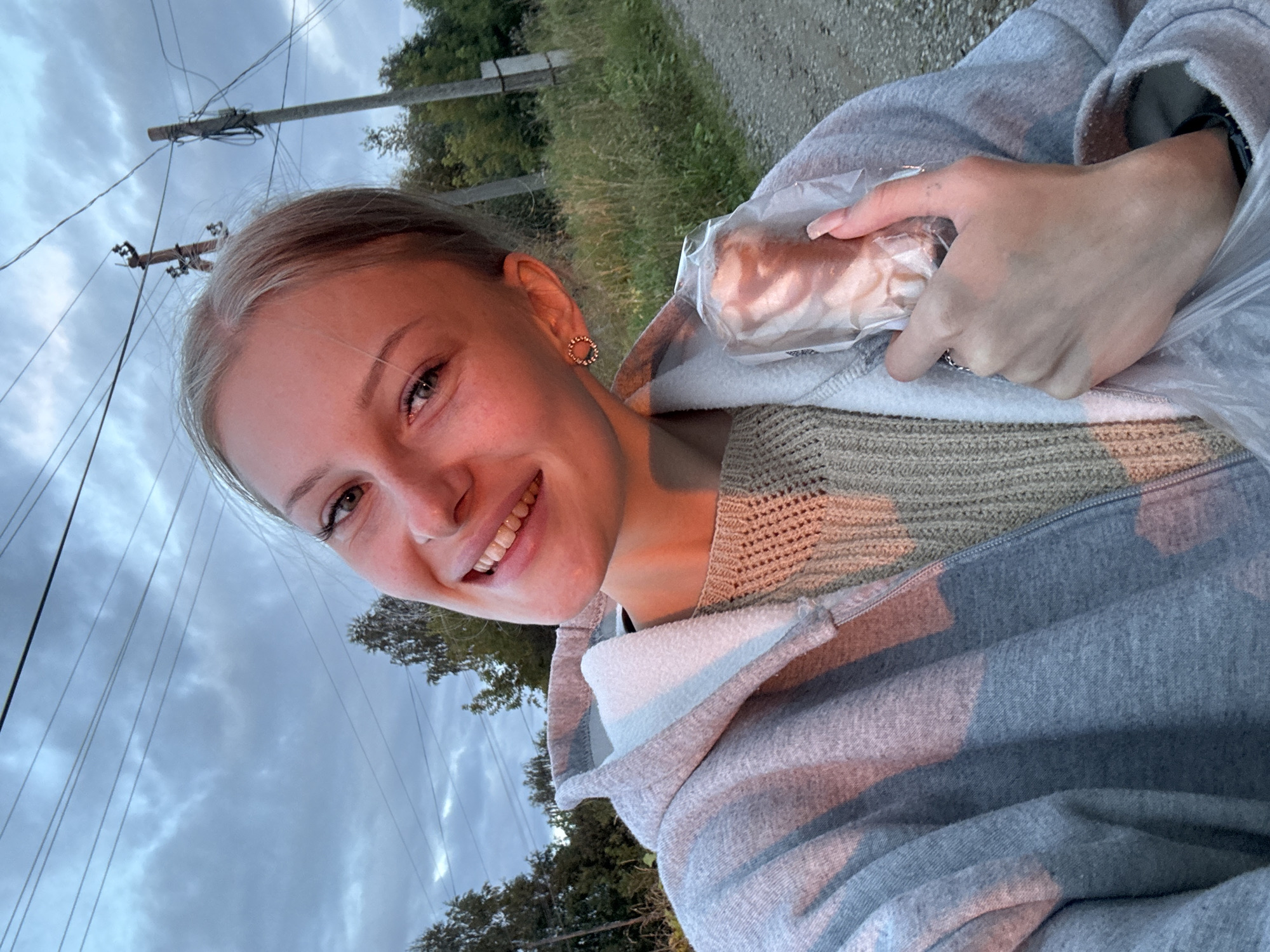 A young Caucasian woman with blonde hair pulled back and fair skin is taking a selfie outdoors. She is smiling and holding a wrapped food item in her right hand. She is wearing a grey hooded sweatshirt over a light brown knitted sweater. In the background, there is a dirt road, tall grass, trees, power lines, and utility poles, suggesting a rural or semi-rural setting. The sky is overcast with clouds. The lighting indicates it may be dusk or dawn. The location is identified as Borodulino, Russia.