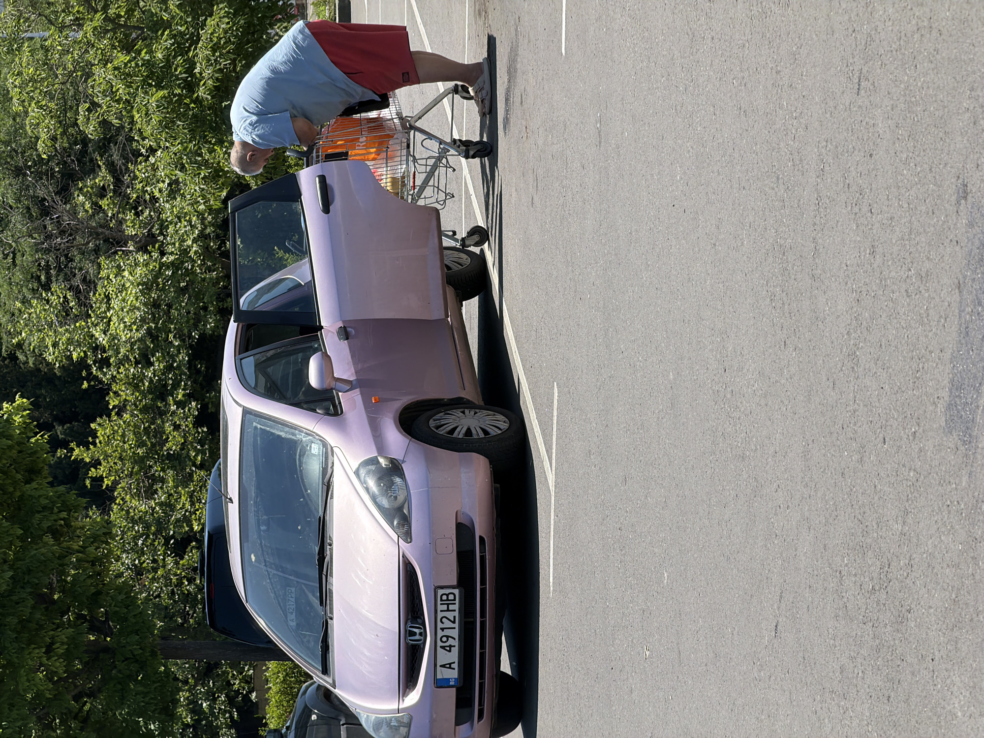 A man in a blue t-shirt and red shorts is loading items from a shopping cart into the open rear passenger door of a light pink Honda Jazz. The car has a license plate that reads "A 4912 HB." The scene takes place in a paved parking lot, likely in Burgas, Bulgaria, given the license plate's format. Lush green trees form the background, suggesting a sunny day. A portion of another dark-colored vehicle is visible to the left of the Honda.