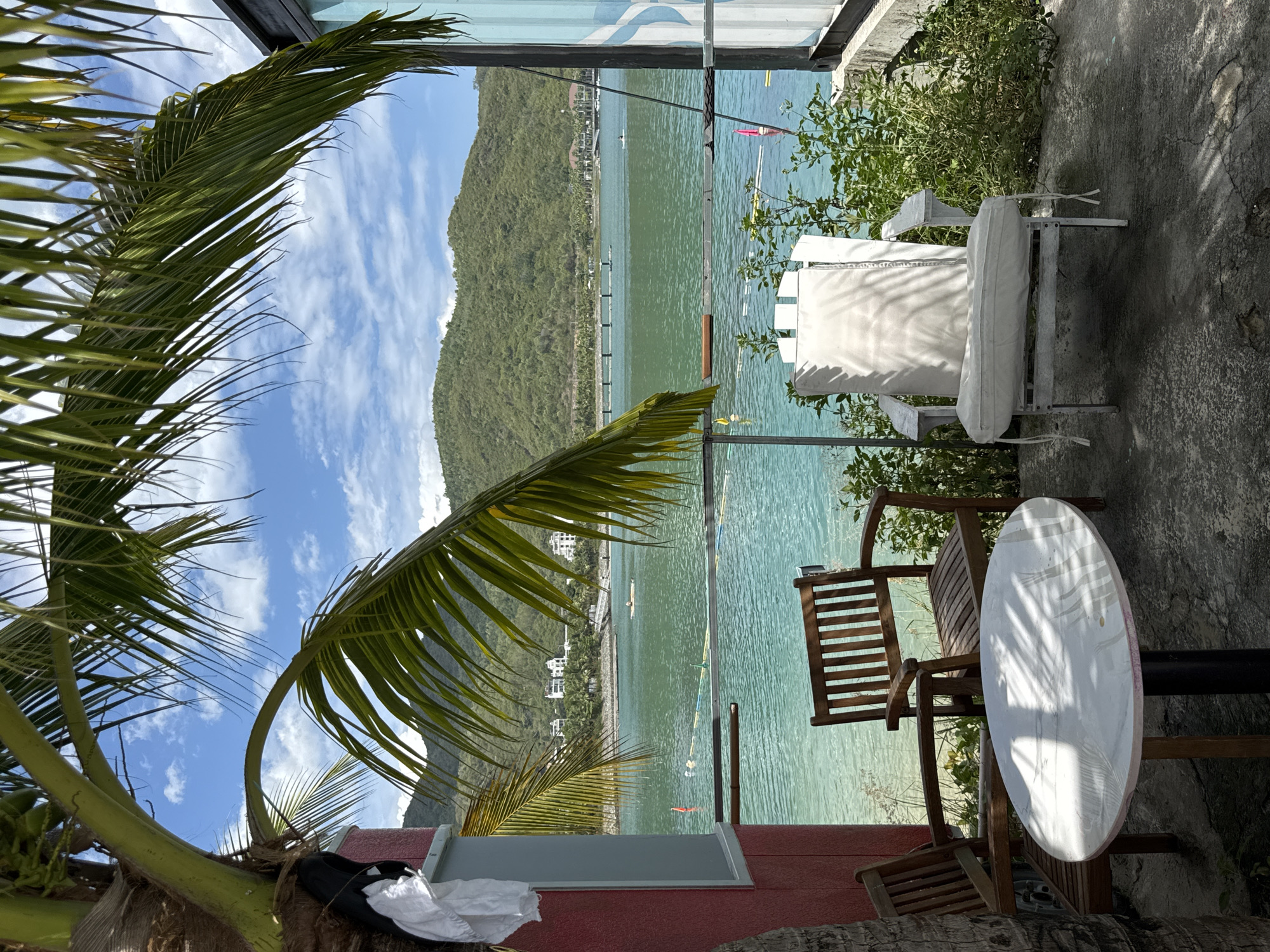 This image depicts an outdoor seating area overlooking a body of water and lush green hills, characteristic of Nha Trang, Vietnam. In the foreground, a small white oval table with a black pedestal base is positioned on a textured concrete patio. Two wooden chairs with slatted backs and seats flank the table. To the right, a white wooden Adirondack-style chair with a white cushion is placed on the patio, with a dense growth of green foliage behind it. A palm tree with large fronds extends into the frame from the left, casting dappled shadows on the table and patio.
Beyond the seating area, the turquoise water of the bay is visible, with a few buoys and possibly a kayak or small boat on the surface. In the middle ground, a row of pilings or a pier stretches across the water, with buildings situated along the shoreline behind it. The background is dominated by rolling green hills covered in vegetation. The sky is blue with scattered white clouds. The overall impression is a serene, tropical setting, likely a resort or restaurant patio in Nha Trang.