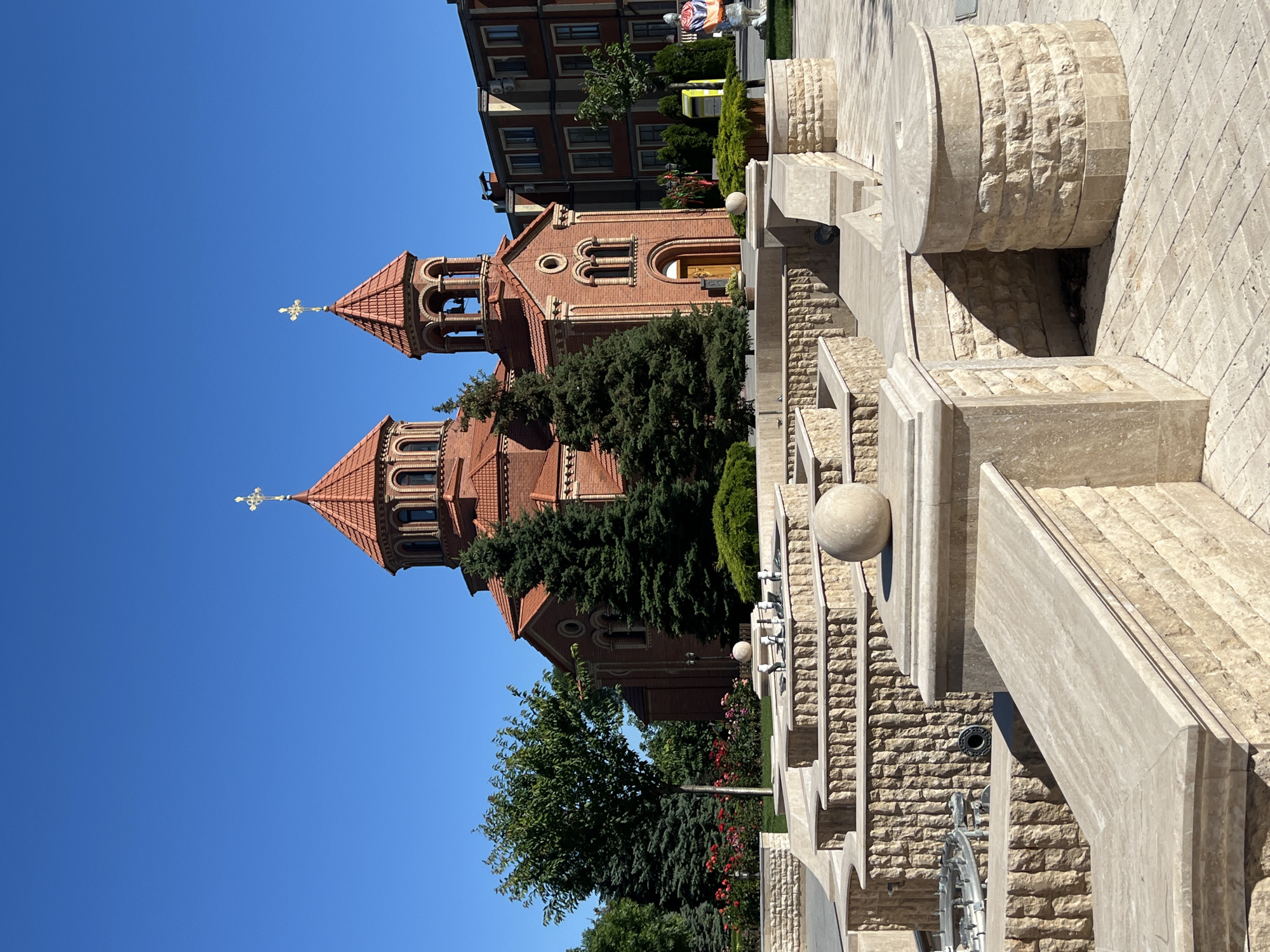 The image shows a brick church with a red tiled roof and two prominent domes, one larger than the other. The larger dome is topped with a golden cross, as is the smaller bell tower. The church has arched windows and a central entrance with a wooden door. In front of the church is a multi-tiered fountain made of light-colored stone, with several tiers of textured stone walls and flat stone surfaces. A spherical stone sits atop one of the fountain's levels. Several evergreen trees and some flowering bushes are visible behind the fountain and to the left of the church. A modern building with multiple windows is located to the right of the church. The sky is clear and blue. The scene appears to be a public space, possibly a park or a square, with paved ground. This is the Armenian Apostolic Church of the Holy Resurrection in Odesa, Ukraine.