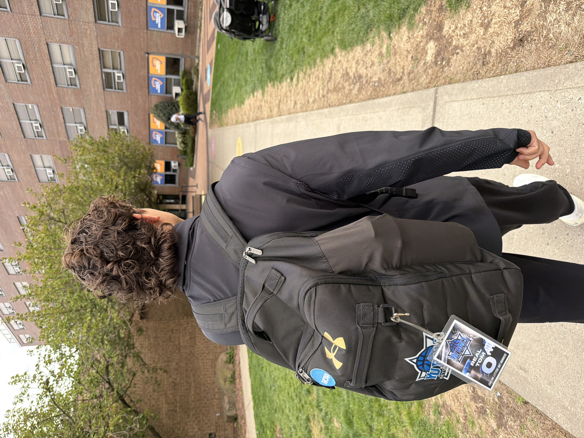 The image captures a person walking outdoors, viewed from behind, along a paved path next to a grassy area. In the background, a multi-story brick building with numerous windows, some featuring air conditioning units, dominates the scene. Several windows display orange and blue banners with the letters "YU" and a logo, suggesting a university setting.
The subject is wearing a dark, long-sleeved top or jacket with a subtle dotted pattern on the sleeves, and dark pants. They have curly, dark hair. A black backpack is carried, prominently featuring a gold Under Armour logo and a circular light blue pin with "NCAA" in white text. Attached to the backpack is a clear ID holder containing a pass.
The pass is basketball-themed and displays the text "YU BALL BEYOND THE COURT," "REAL TOM O," and "CHASING LETTERS NOT NUMBERS." Another logo on the backpack, adjacent to the pass, also shows a stylized "YU" with a basketball design.
The scene suggests a daytime setting with overcast weather, providing even lighting. The presence of "YU" branding on both the building and the person's accessories, combined with the "NCAA" pin, strongly indicates a collegiate environment, likely Yeshiva University in New York, United States of America, given the location context. The person appears to be moving across the campus, possibly to or from a sports-related event.