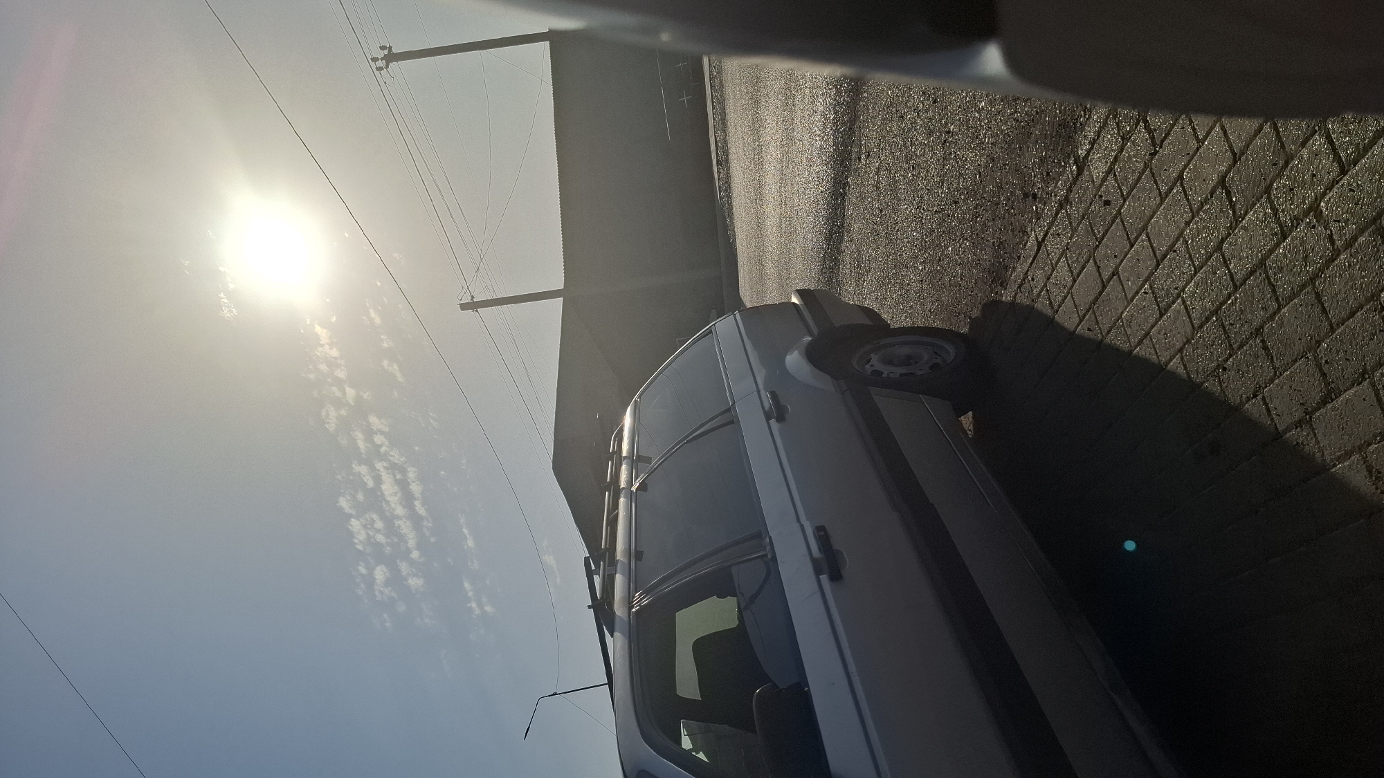 The image shows a silver station wagon parked on a paved area, possibly a street or parking lot, in Шұбарсу, Kazakhstan. The sun is bright and high in the sky, casting long shadows. The car is the primary subject, viewed from the side. In the background, there is a dark, low-rise building or wall, and power lines stretch across the frame. The ground is a mix of asphalt and brick paving stones. There are no people visible in the image. The overall impression is a sunny day in an urban or semi-urban environment.