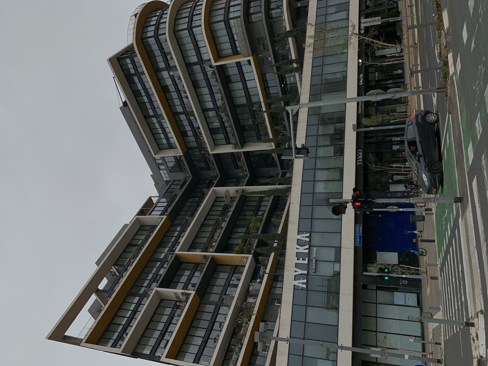 The image shows a modern, multi-story building in Tel-Aviv, Israel. The building, named "AYEKA", has a unique architectural design with angled balconies. Adjacent to the building, a street is visible, including a car stopped at a red traffic light, pedestrian crosswalks, and a bike lane. Storefronts of businesses named "TIAGO" and "GALITA" can be seen along the building's ground level. The sky is overcast.