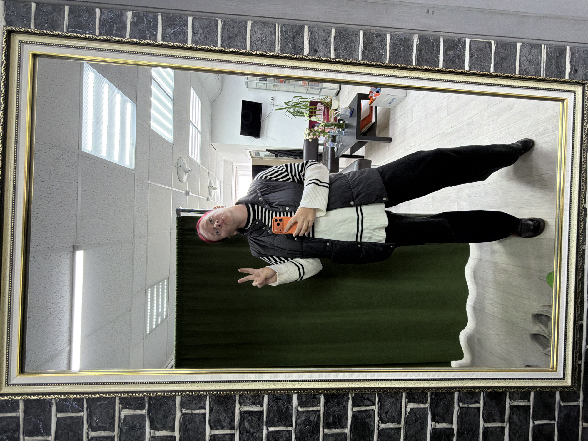 A person with pink hair is standing in front of a mirror, holding an orange cell phone and making a peace sign with their left hand. They are wearing a black puffer vest over a white and black striped long-sleeved shirt, and black pants. The mirror is framed with a gold and black textured border. Behind the person, a green curtain is visible, and in the reflection of the mirror, a salon-like environment can be seen with a black coffee table, chairs, plants, and a television mounted on the wall. The location is the city of Chaikovsky, Russia, specifically within a commercial establishment that appears to be a salon or similar business, characterized by the furnishings and the overall interior design. The lighting suggests indoor fluorescent lighting, typical of such spaces. The floor is a light-colored laminate.