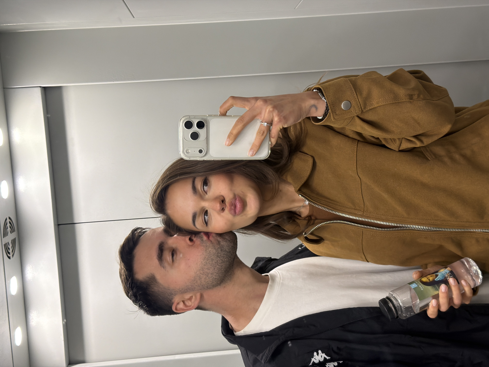 An intimate selfie shows a couple posing playfully in an indoor setting, likely an elevator or a mirrored room, with the image rotated 90 degrees counter-clockwise.
The woman, positioned in the upper portion of the rotated image, is taking the photo. She has medium-brown hair and is wearing a brown jacket with a silver zipper. Her right hand, adorned with a white ring on her ring finger, holds a light-colored smartphone with a clear case and a triple camera lens system. A small crescent moon tattoo is visible on her right inner wrist. She makes a "kissing face" towards the camera.
Below her, in the lower portion of the rotated image, the man kisses her cheek or temple. He has short dark hair and a beard, and wears a white t-shirt beneath a black jacket with a white logo (resembling the Kappa brand) on the left side. In his left hand, he holds a small, clear plastic water bottle with a black cap. The bottle's label features an image of a classical painting, likely the Mona Lisa, and some text.
The background consists of smooth, light gray or white walls. On what appears to be the left side of the room (bottom in the original orientation), vertical strip lights and a vent are visible, suggesting bright artificial lighting. The overall mood is affectionate and lighthearted.