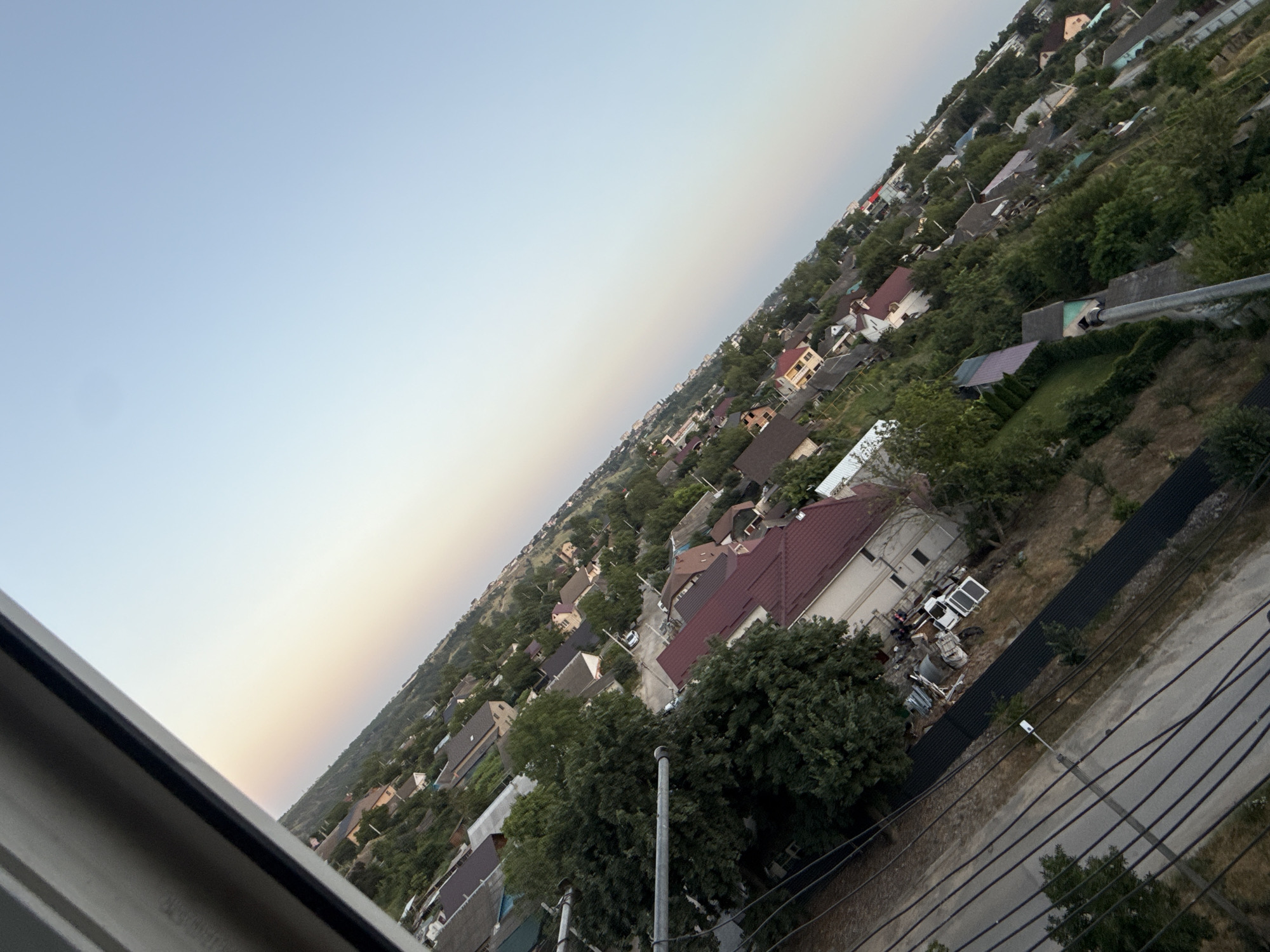 The image shows an aerial view of a residential area in Cahul, Moldova, characterized by a dense collection of houses with various roof colors, predominantly red and gray. The houses are nestled among numerous green trees, creating a lush, suburban landscape. The perspective is angled, with the sky dominating the upper right portion of the frame. A subtle gradient of colors, from a pale yellow-orange near the horizon to a clear blue, suggests either sunrise or sunset. In the foreground, a metal pole and power lines are visible, indicating the presence of infrastructure within the neighborhood. The overall scene depicts a quiet, possibly rural or semi-urban, residential setting during a transitional period of daylight.