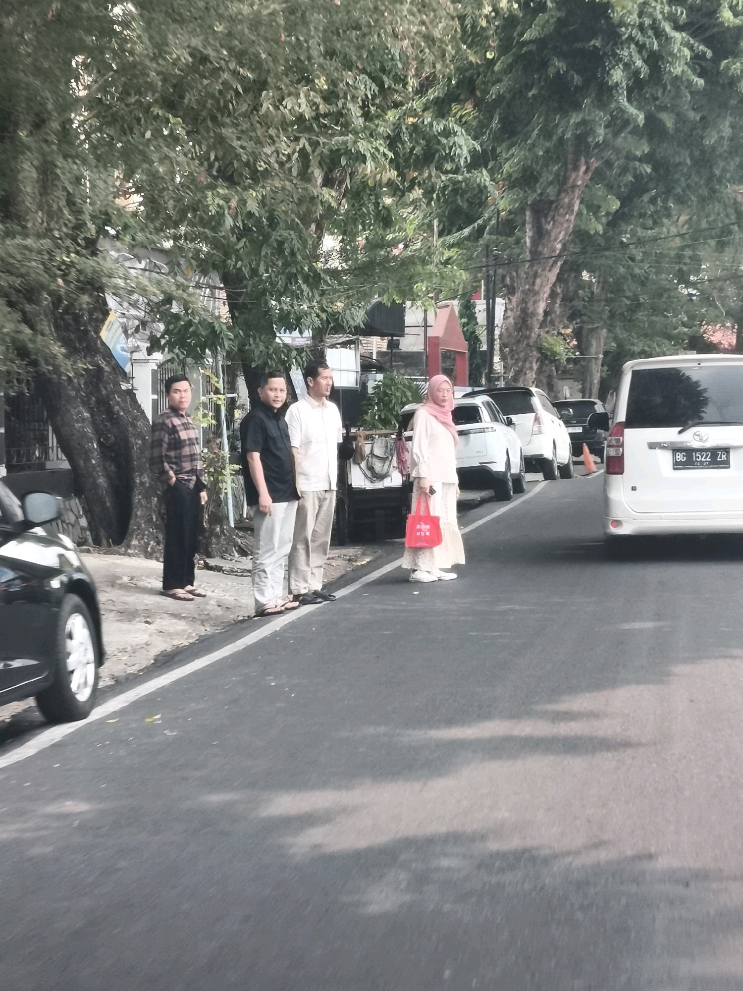 The image captures a street scene in Palembang, Indonesia, with several individuals standing along the roadside. In the foreground, a black car is partially visible on the left. Further down the road, three men are standing in a line, dressed in casual attire. The man on the left wears a plaid shirt and dark pants, the middle man is in a black shirt and light pants, and the man on the right is in a light-colored shirt and light pants. To their right, a woman is standing, wearing a pink hijab and a light-colored dress, carrying a red bag. Behind the group, several cars are parked or driving, including a white van with the license plate BG 1522 ZR on the right side of the frame. The environment is characterized by trees lining the street and buildings in the background, suggesting an urban or suburban setting. The lighting indicates it might be daytime, possibly late afternoon due to the shadows.