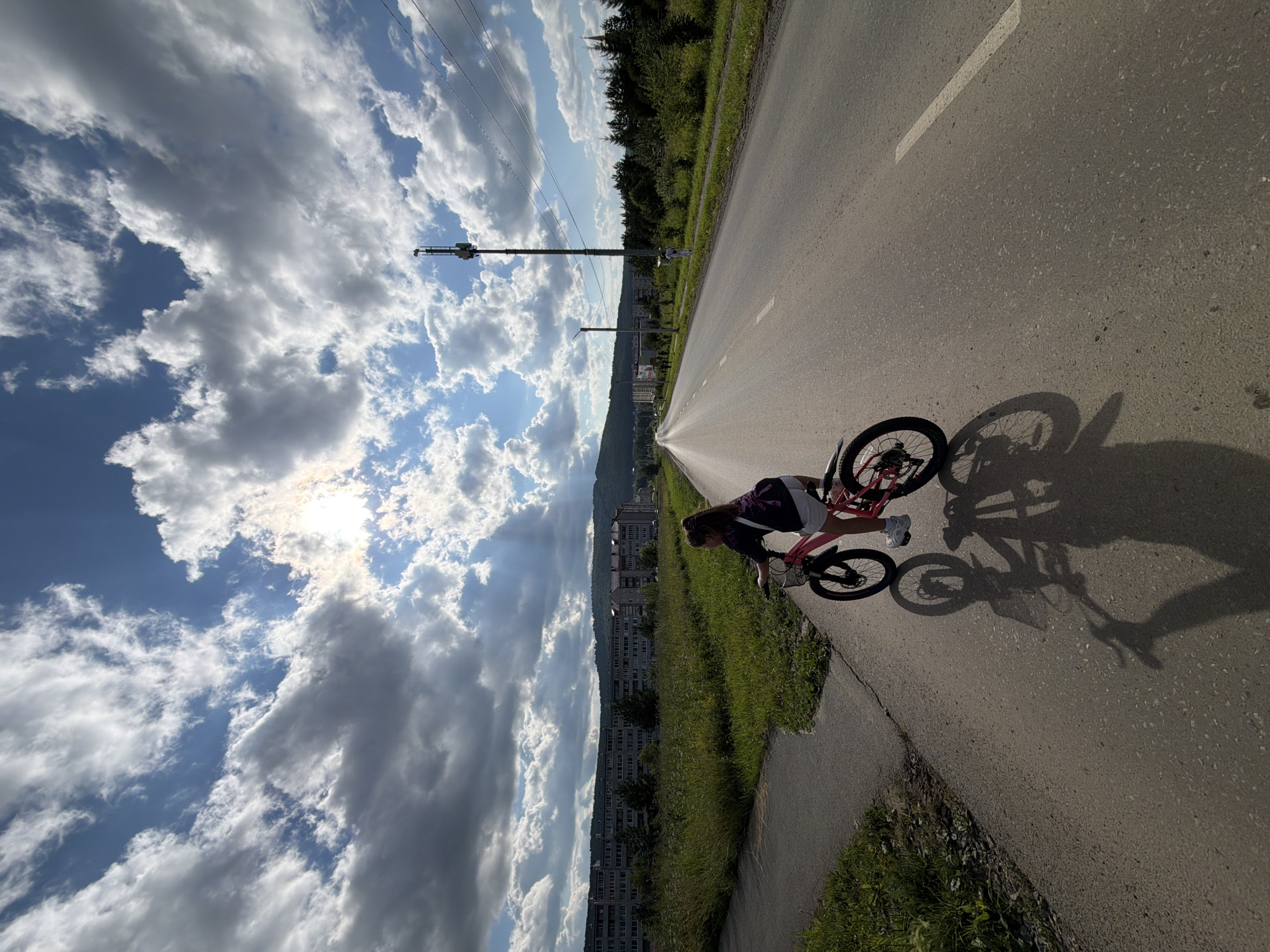 A young girl with braided brown hair, wearing a dark t-shirt and white shorts, is riding a pink bicycle on an asphalt road in an unknown city. She is facing away from the camera, towards the right side of the frame. The bicycle has black wheels and a basket on the handlebars. A dark backpack is strapped to the back of the bicycle. The sun is shining brightly through a dramatic sky filled with large, white and grey clouds, casting long shadows of the girl and her bicycle onto the road.
In the background, to the left, is a long, multi-story residential building with many windows, partially obscured by tall green grass and trees. Beyond the building, a range of tree-covered hills is visible. To the right of the road, there is a sidewalk, followed by more grass, trees, and a tall communication tower with multiple antennas. Power lines run overhead from the tower. The road stretches into the distance, with a white line marking the center. The overall scene suggests a bright, sunny day in a semi-urban or suburban environment.
