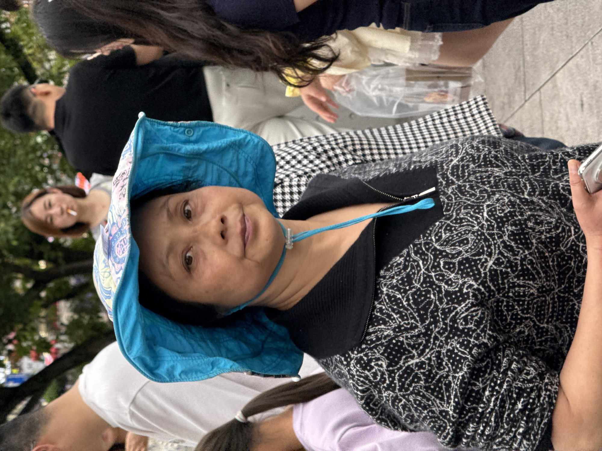 The image shows a woman wearing a light blue sun hat with a drawstring around her neck. She is wearing a black zip-up jacket over a black top, with the jacket having a white-patterned design. Her skin tone appears to be Asian.
Behind the woman are several other people, seemingly in a public area. One person is wearing a black shirt, another a white shirt. A woman further back is holding a cigarette. There is a girl in the foreground wearing a lavender shirt with long hair in a pony tail. In the background is greenery, potentially trees.
The image was taken in an outdoor environment, likely a crowded area. It's unclear where specifically the image was taken in Yuzhong District, China.