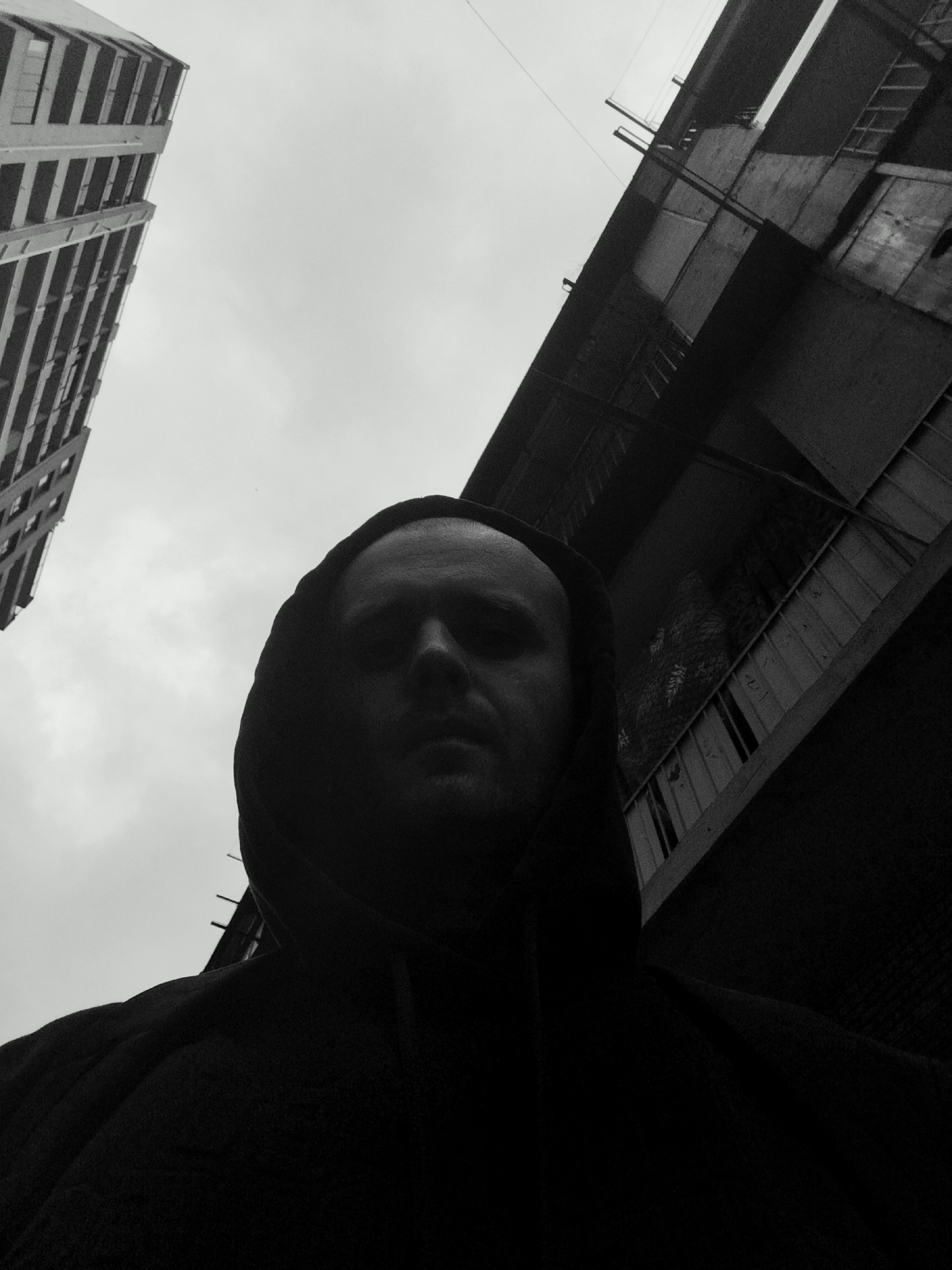 A low-angle, black and white, outdoor, medium shot shows the head and shoulders of a person wearing a dark hoodie. The person's face is mostly obscured by shadow, with only the forehead and eyes faintly visible. The background is dominated by the overcast sky and the angular architecture of two buildings. On the left, a tall, rectangular building with many windows is visible, angled upwards. To the right, a darker, more textured building with visible balconies and structural elements extends upwards, also angled. The scene appears to be in an urban environment. While the image does not explicitly show details specific to Batumi, Georgia, the architectural style is consistent with some areas of the city.