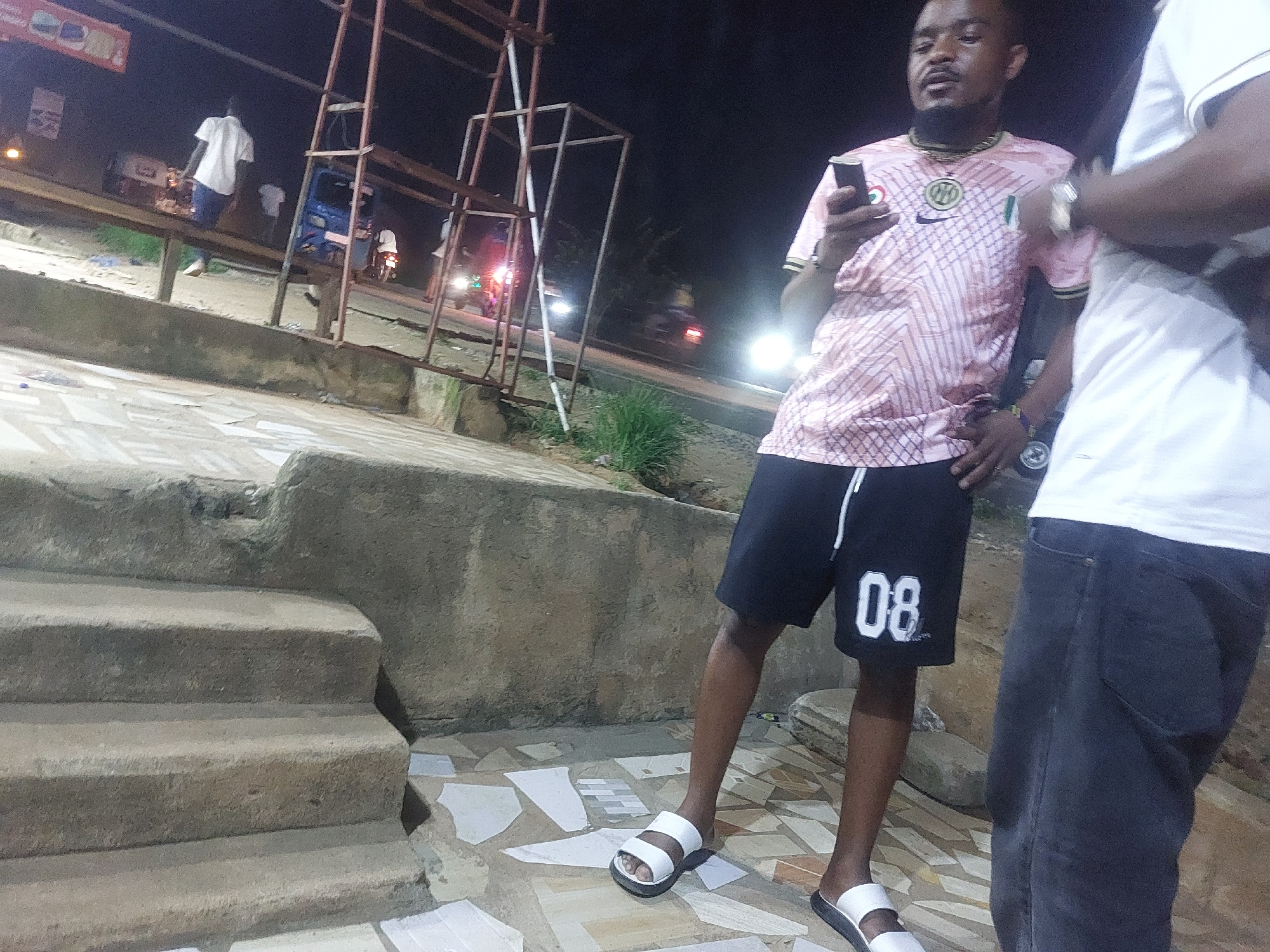 A nighttime shot depicts a man holding a mobile phone. He is wearing a pink patterned shirt, black shorts with the number 08 on them, and white sandals. He stands on a tiled surface in front of a concrete structure with steps. Another person is partially visible on the right side of the frame, wearing a white shirt and blue jeans. In the background, blurred lights suggest street activity, including figures on a bench, a vehicle, and other unidentified structures. The setting appears to be an urban environment, consistent with an area in Dar es-Salaam, Tanzania.