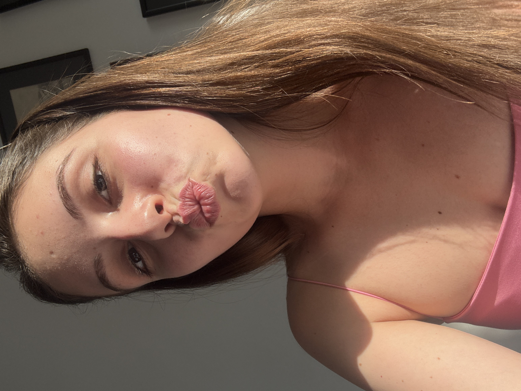 A young woman with long brown hair is pictured in a close-up, selfie-style shot. She is wearing a pink spaghetti strap top and is puckering her lips. Sunlight casts shadows across her face and shoulder. The background is a plain gray wall, with two framed pictures visible on the right side of the image. The context provided about Burgas, Bulgaria, is not visually evident in the image itself.