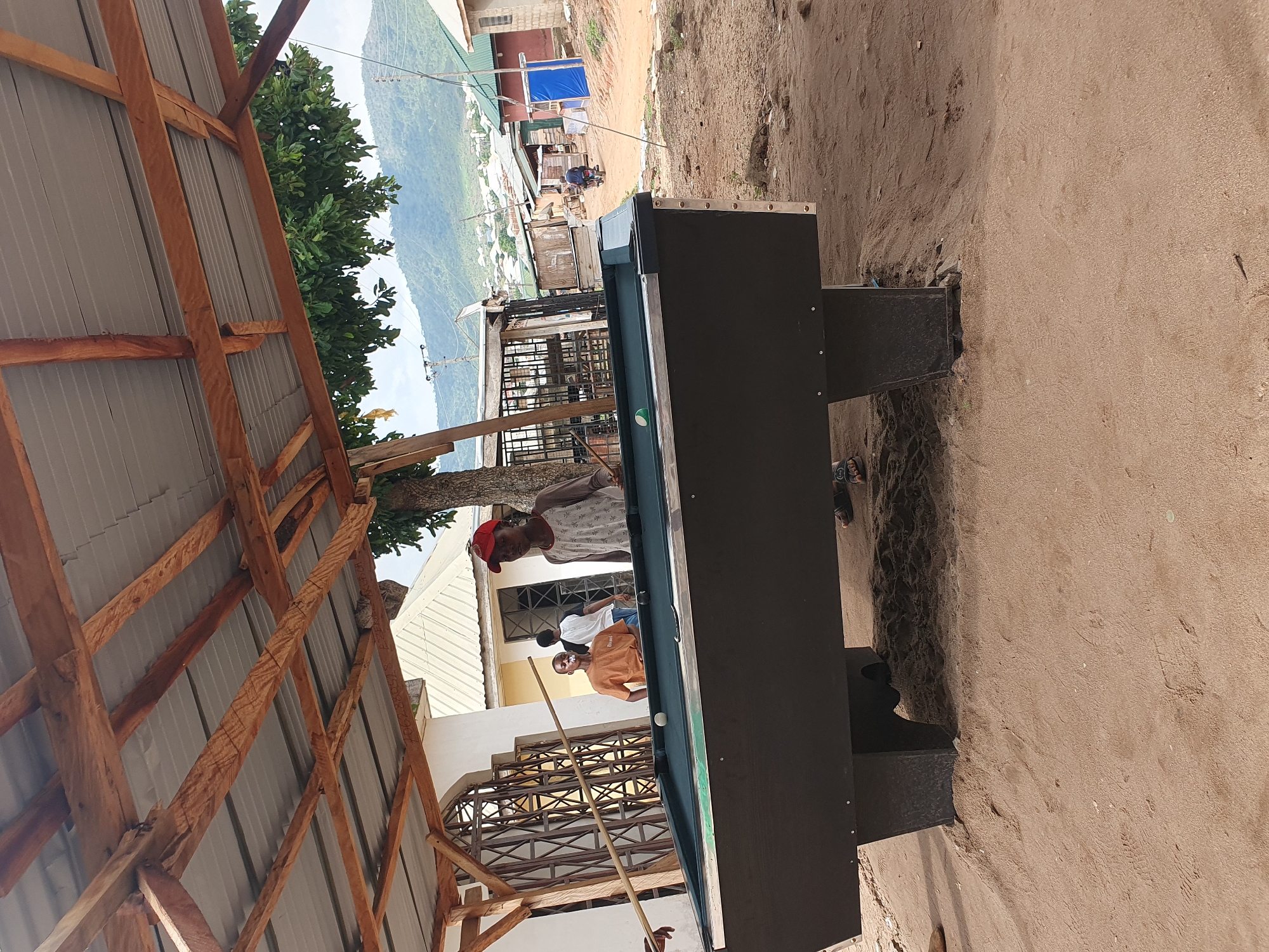 A slightly high-angle outdoor shot depicts a black pool table on a sandy or dirt ground, with three people playing pool or socializing around it. The players are of African descent. One person is wearing an orange shirt, another a white shirt, and the third is wearing a red cap and patterned shirt. The foreground includes the wooden frame of a corrugated metal roof overhanging the scene. In the background, there is a glimpse of a residential area with houses and trees. In the distance, mountains are visible. The photo appears to be taken in Lokoja, Nigeria, based on the typical environment.