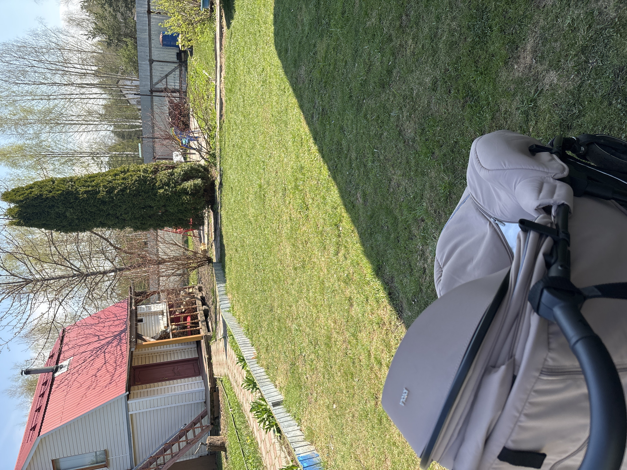 The image shows a residential scene in Мещёры, Russia. A beige stroller occupies the foreground, partially obscuring the view. Beyond the stroller is a lawn, with varying shades of green suggesting sun and shadow. To the left, a small, white house with a red roof is visible. A wooden structure, possibly a lean-to or firewood storage, is attached to the house. In the background, trees and a fence line are present, indicating the edge of the property. The sky is visible through the bare branches of the trees.