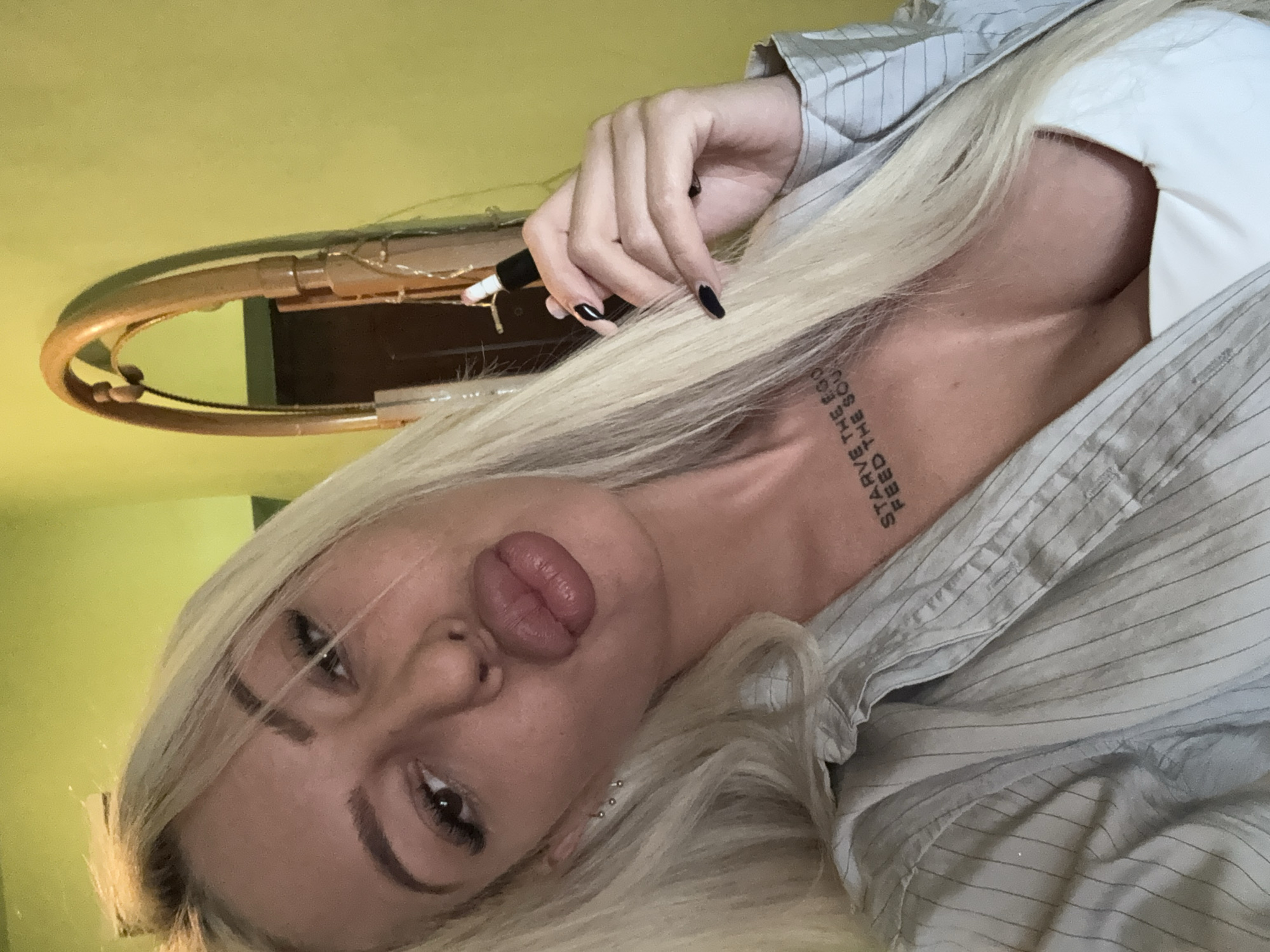 A woman with platinum blonde hair and black nail polish is holding a pen in her hand. She has a tattoo on her chest that reads "Starve the Ego, Feed the Soul". She is wearing a light blue pinstripe shirt. Behind her is a wall with an oval mirror. The location is in Plovdiv, Bulgaria.