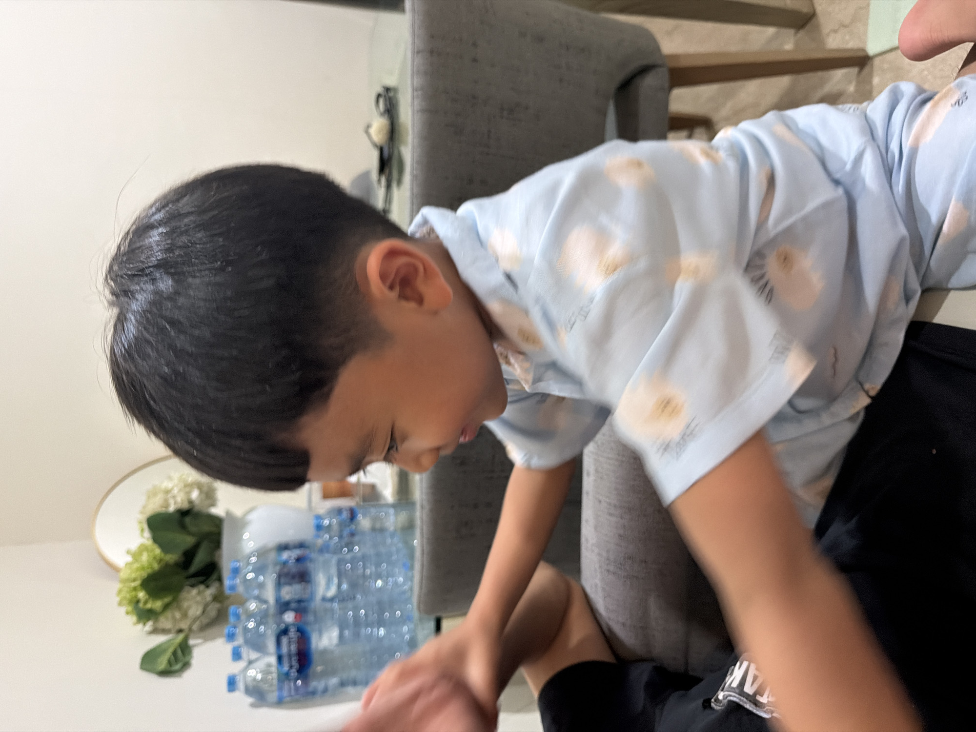 The image depicts a young boy in a light blue collared shirt with a floral pattern, sitting indoors and interacting with another person whose arm is partially visible. The boy is looking to his right and smiling, with his right arm extended and bent at the elbow. Behind him, several bottles of water are lined up on a surface, and in the background, there is a vase of flowers, a round mirror, and a dining table with chairs. The overall setting suggests a domestic environment within a city. Based on the provided location of Central Jakarta, Indonesia, this scene likely takes place in a residential or commercial establishment in that area.
