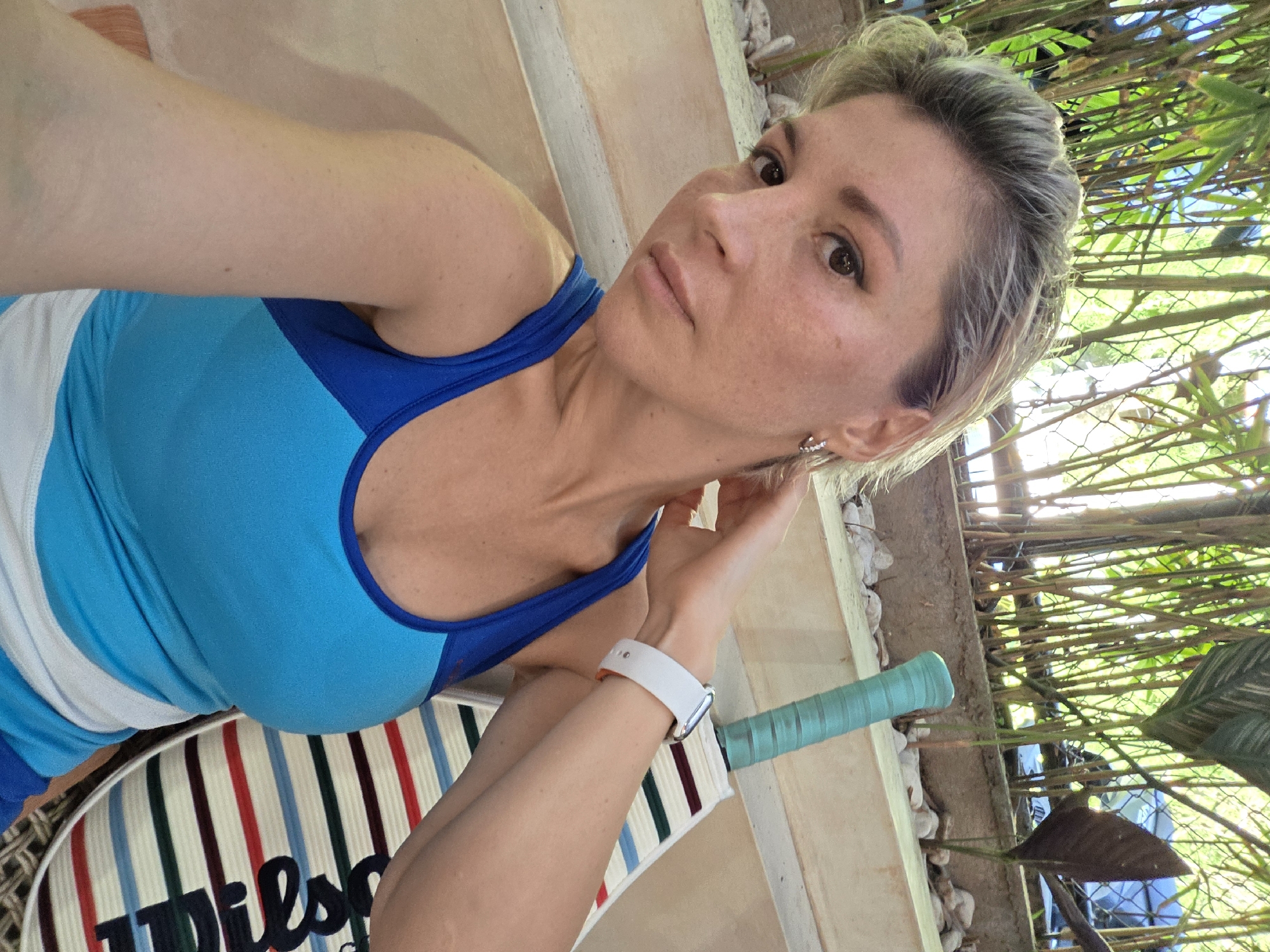 A woman is captured in a selfie, seated outdoors with a tropical garden and bamboo stalks in the background. She is wearing a blue and white athletic tank top and a white smartwatch. To her right, a tennis racket with a light blue grip rests on a striped cushion. The scene appears to be in North Kuta, Indonesia, judging by the lush vegetation and the warm, natural lighting.
