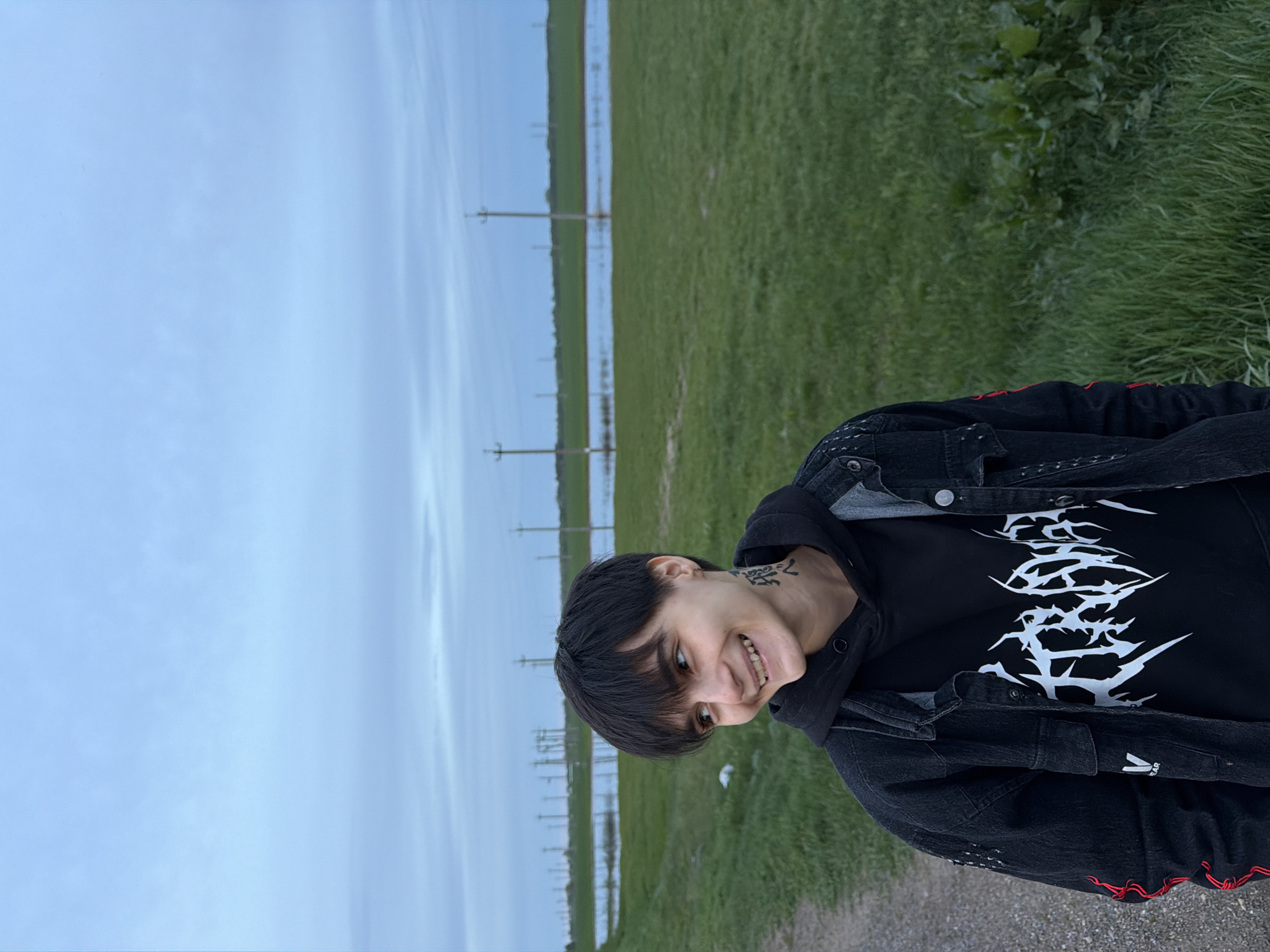 A photo taken in Azyakul', Russia, depicts a person standing in a grassy area. The sky is overcast. Power lines are visible in the background. The person is wearing a black hoodie with a white graphic, and a black denim jacket with red stitching. They have short, dark hair and a tattoo on their neck. They are smiling.
