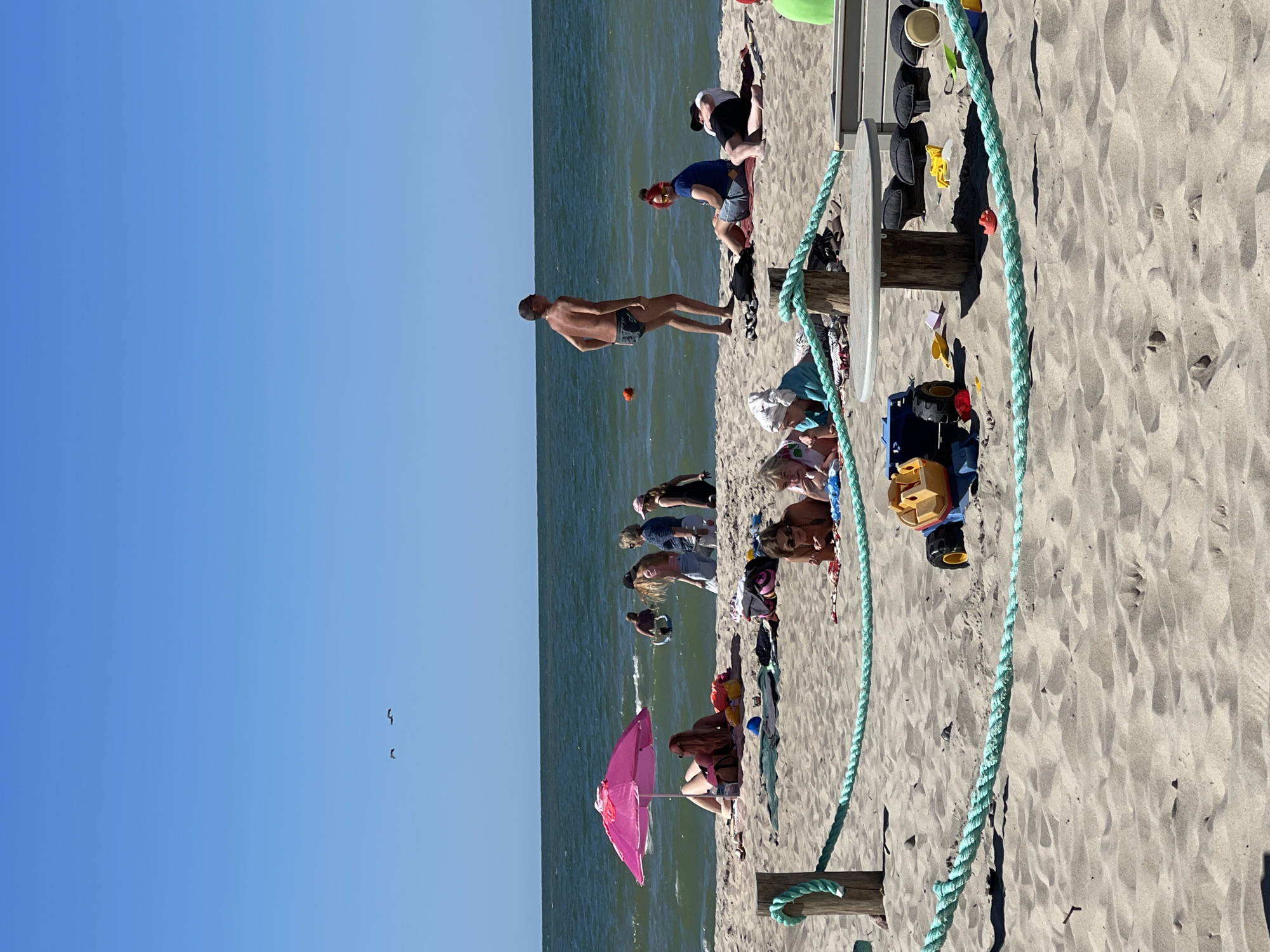 The image depicts a sunny day at the beach in Zelenogradsk, Russia. A group of people are enjoying themselves on the sandy shore, with the calm, blue ocean stretching out behind them. The water appears to be relatively shallow near the shore, and some individuals are wading or standing in it.
On the sand, several people are relaxing, sunbathing, or playing. A man in swim trunks stands near the water's edge, facing away from the camera. Further up the beach, a pink umbrella provides shade for someone sitting on the sand. Children's toys, including a blue truck and a red toy, are scattered on the sand, suggesting a family-friendly atmosphere.
A wooden structure with a light-colored top, possibly a picnic table or a bench, is visible, with some personal belongings like hats and bags placed on it. A thick, turquoise rope is strung across the foreground, adding a nautical or recreational element to the scene. Two birds are visible flying in the clear blue sky. The lighting suggests it is daytime, likely mid-morning or afternoon, with bright sunlight casting shadows on the sand. The overall mood appears relaxed and leisurely, indicative of a typical beach day. No visible text is present in the image.