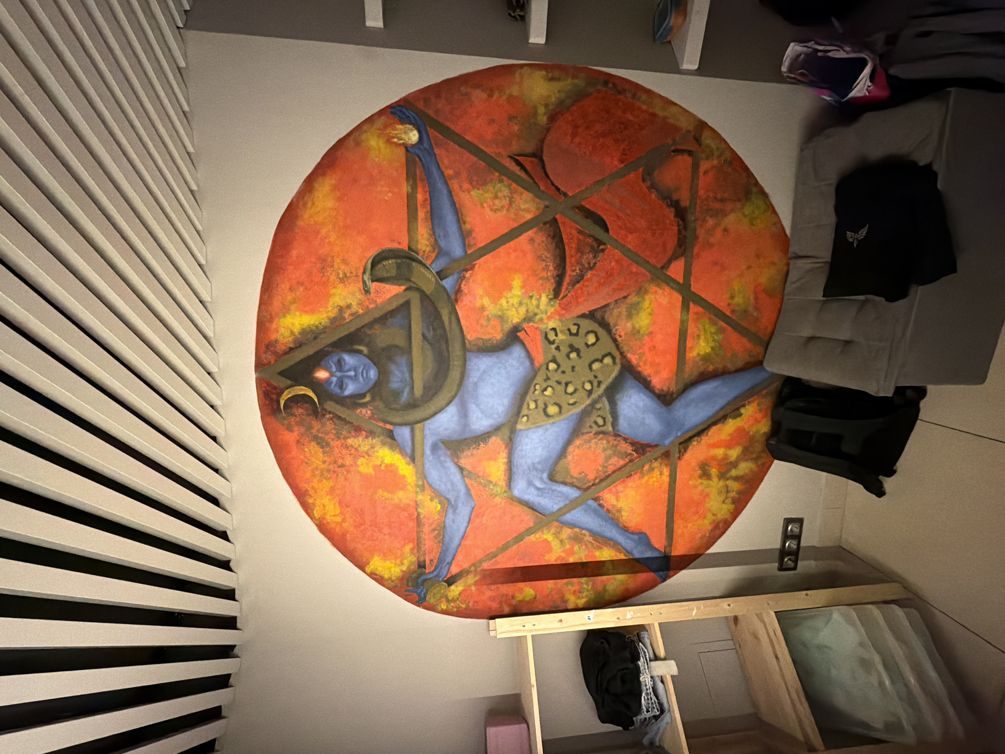 The image shows a wall with a large, circular painting. The painting depicts a blue-skinned figure with a headdress, set against a backdrop of orange and yellow hues. The figure appears to be in a dynamic pose, with what seems to be a leopard-print garment. Wooden slats are visible on the left side of the image, possibly indicating a wall paneling or partition. In the lower part of the frame, there's a wooden structure, possibly a storage unit or shelving. There are various objects scattered around, including a black bag, a gray cushion, and some other items, suggesting an indoor space. The image does not provide enough detail to determine the specific location in Moscow, Russia or any information about people, actions, or interactions related to the scene.