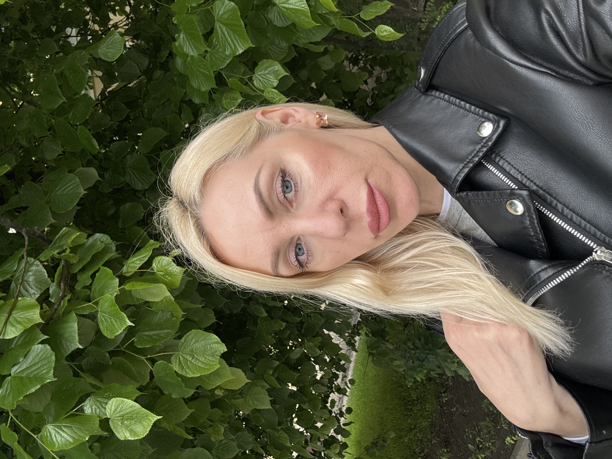 A woman with blonde hair and blue eyes poses in front of a dense green leafy tree in what appears to be a city setting. She is wearing a black leather jacket, and her left arm is partially visible. She has a blush on her cheeks and is wearing lipstick. A shiny square earring adorns her ear. The background shows a glimpse of grass and light, hinting at an urban park or garden-like area within a city.