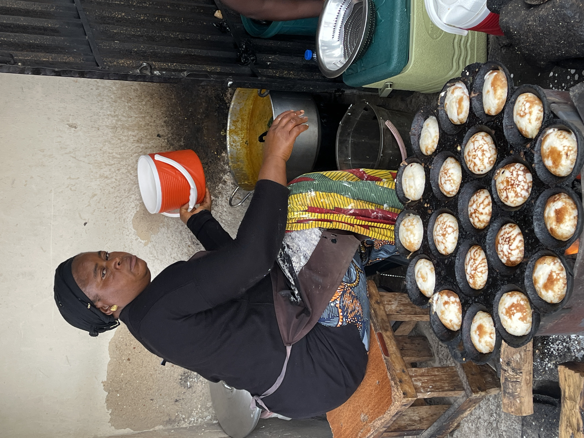 The image depicts an outdoor food stall scene in the city of Jos, Nigeria. A woman, wearing a black headscarf and a black long-sleeved shirt, sits on a wooden stool and is actively preparing food. She is holding an orange and white plastic bucket in her right hand. Her attire includes a patterned apron over a dark garment, and she wears gold earrings. In front of her, a specialized cooking pan with multiple round indentations is placed over an open fire, containing several small, cooked pancake-like items, some of which show browning. To the right of the cooking pan, a large pot filled with a yellow, stew-like substance is visible. Further to the right, a green cooler box and a metal colander are present. In the background, a rustic wall and a dark metal gate are observed. The ground is concrete, with some scattered debris. The overall impression is that of a bustling street food vendor in an urban Nigerian setting.