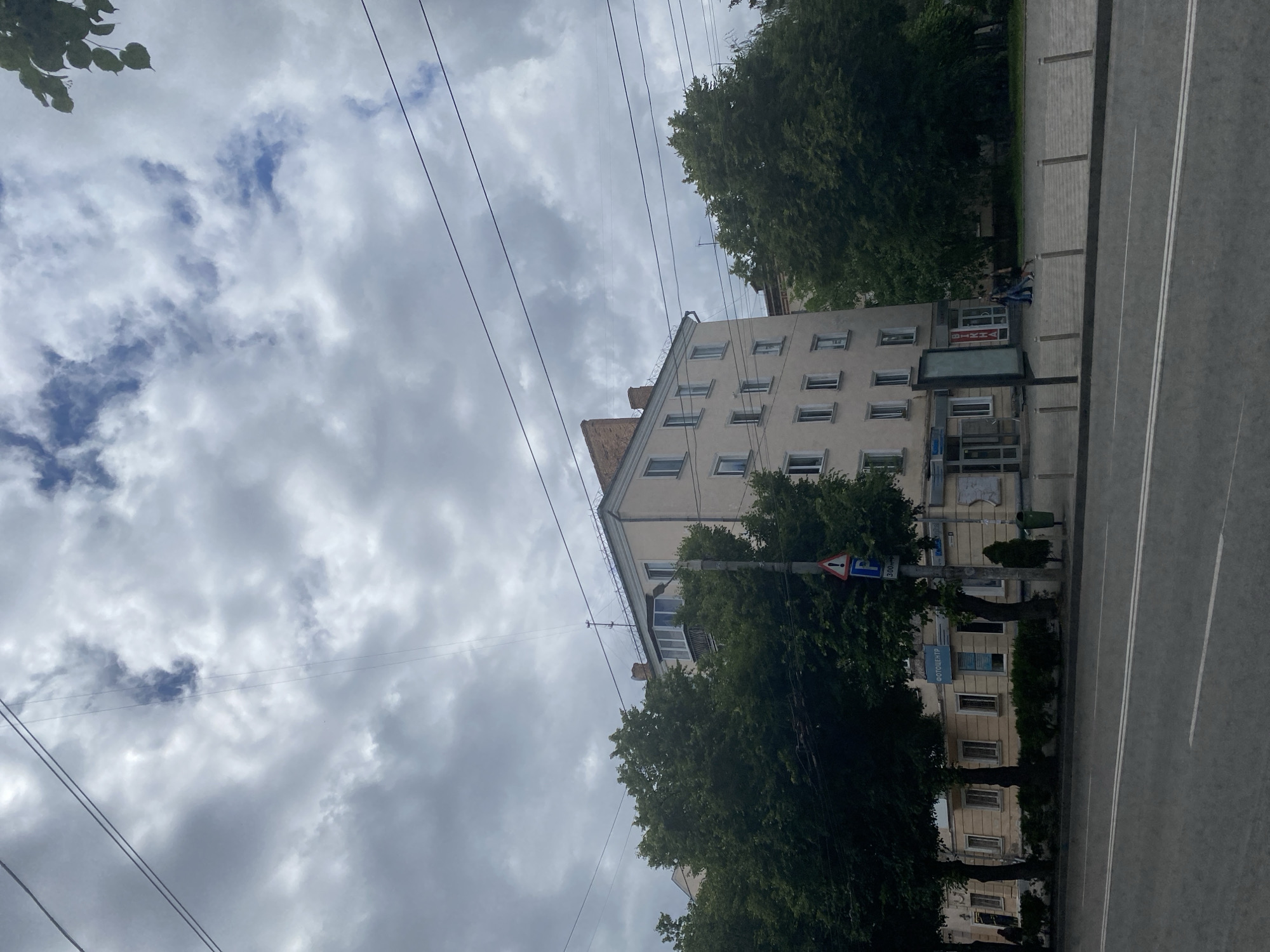 The image shows a street view in an urban environment under a cloudy sky. In the foreground, a gray asphalt road with white lane markings stretches across the bottom of the frame. Behind the road, a sidewalk area is visible, lined with bollards. Adjacent to the sidewalk is a row of trees with dense green foliage. Beyond the trees, a multi-story building with a light-colored facade and numerous windows stands tall. The building has a brick chimney visible at the top. There are also several utility poles and wires crisscrossing the sky and obstructing the view of the building. On the sidewalk, a few individuals are walking. One sign on a pole indicates parking and a distance of 300 meters, and another sign has the word "ФОТОЦЕНТР" (photo center) in blue letters. The overall impression is a typical city street scene on an overcast day.