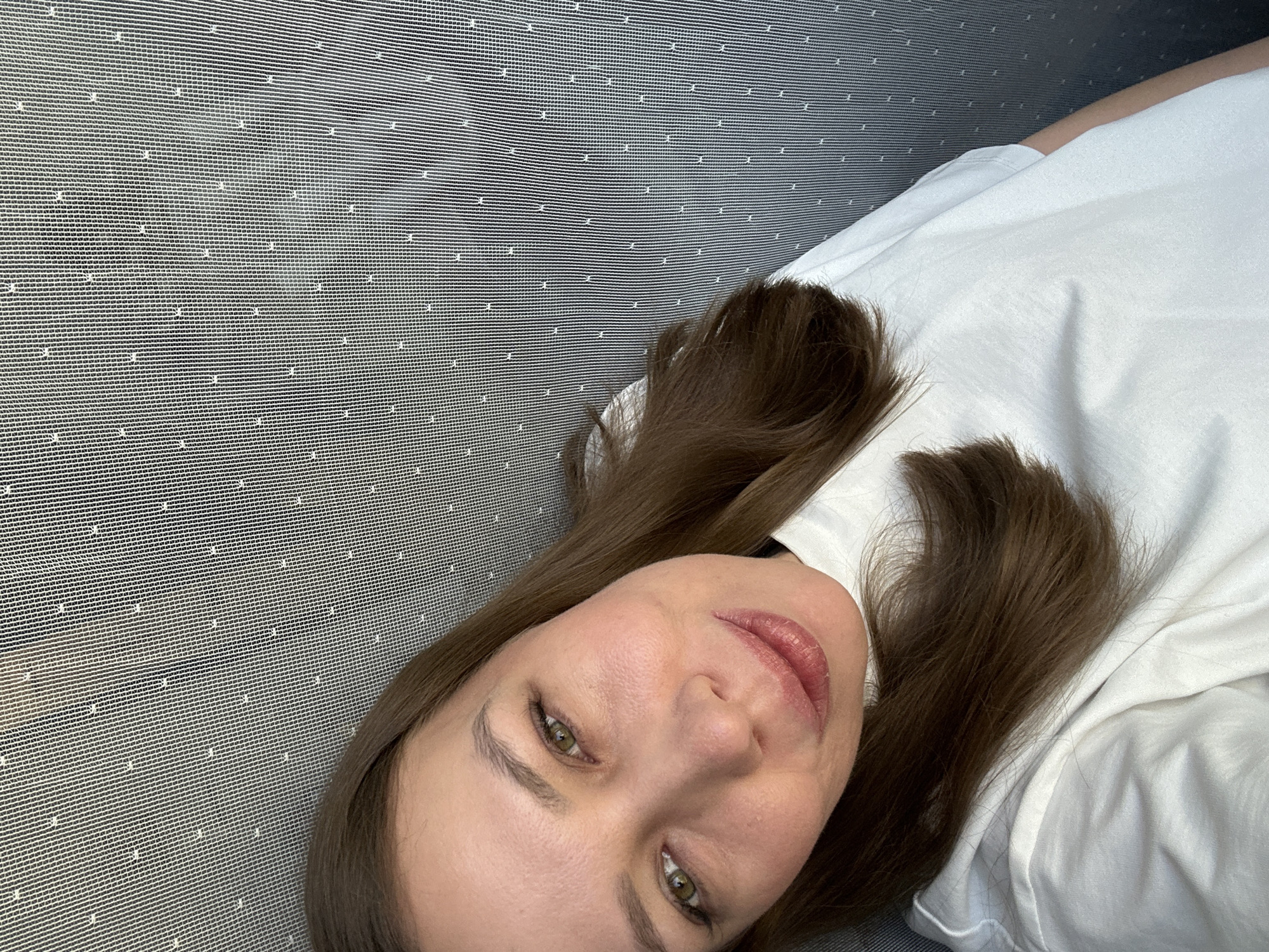 A woman with fair skin, green eyes, and medium-brown hair lies on a gray surface with a pattern of small, white dots. She is wearing a white shirt. The background is a repetitive pattern, seemingly a textured surface or material. The location, Tomilino, Russia, cannot be confirmed from the image alone. The information is insufficient to confirm the city.