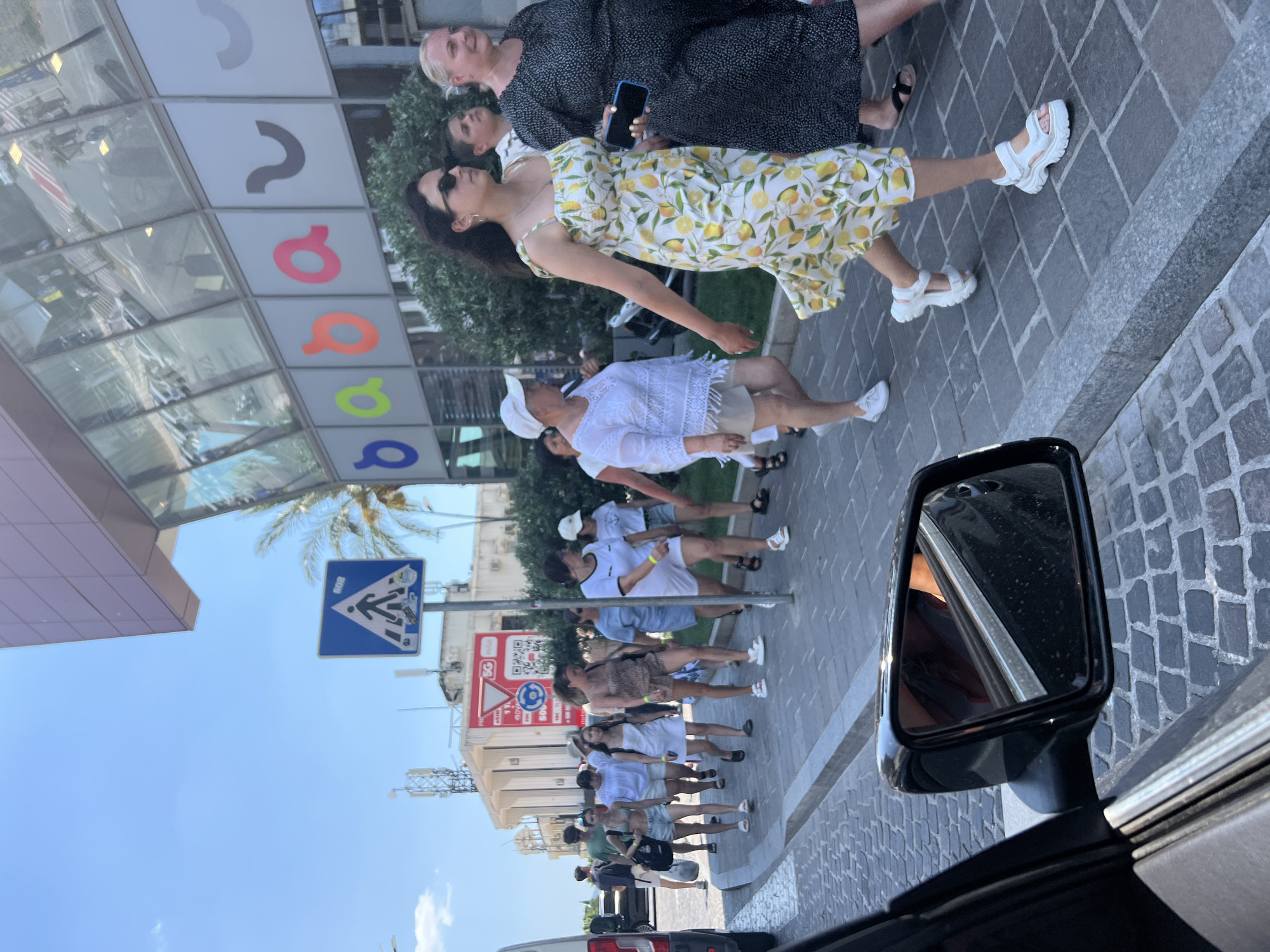 The image captures a street scene in Budva, Montenegro, on a sunny day. A group of people are walking on a cobblestone sidewalk adjacent to a modern building with large glass windows displaying colorful lettering. In the foreground, the side mirror of a black car is visible, reflecting part of the car's interior. A pedestrian crossing sign with a silhouette of a person walking and suitcases is mounted on a pole to the left. The people in the group are dressed in casual summer clothing. One woman in the foreground wears a yellow dress with a lemon print and white sandals. To her right, another woman in a black polka-dot dress walks with a phone in her hand. A young boy with dark hair is seen between them. Further down the sidewalk, a group of younger individuals, primarily women, are walking in the same direction. The building behind them appears to be a commercial establishment, with reflections of traffic and passersby on its facade. The sky is clear and blue.