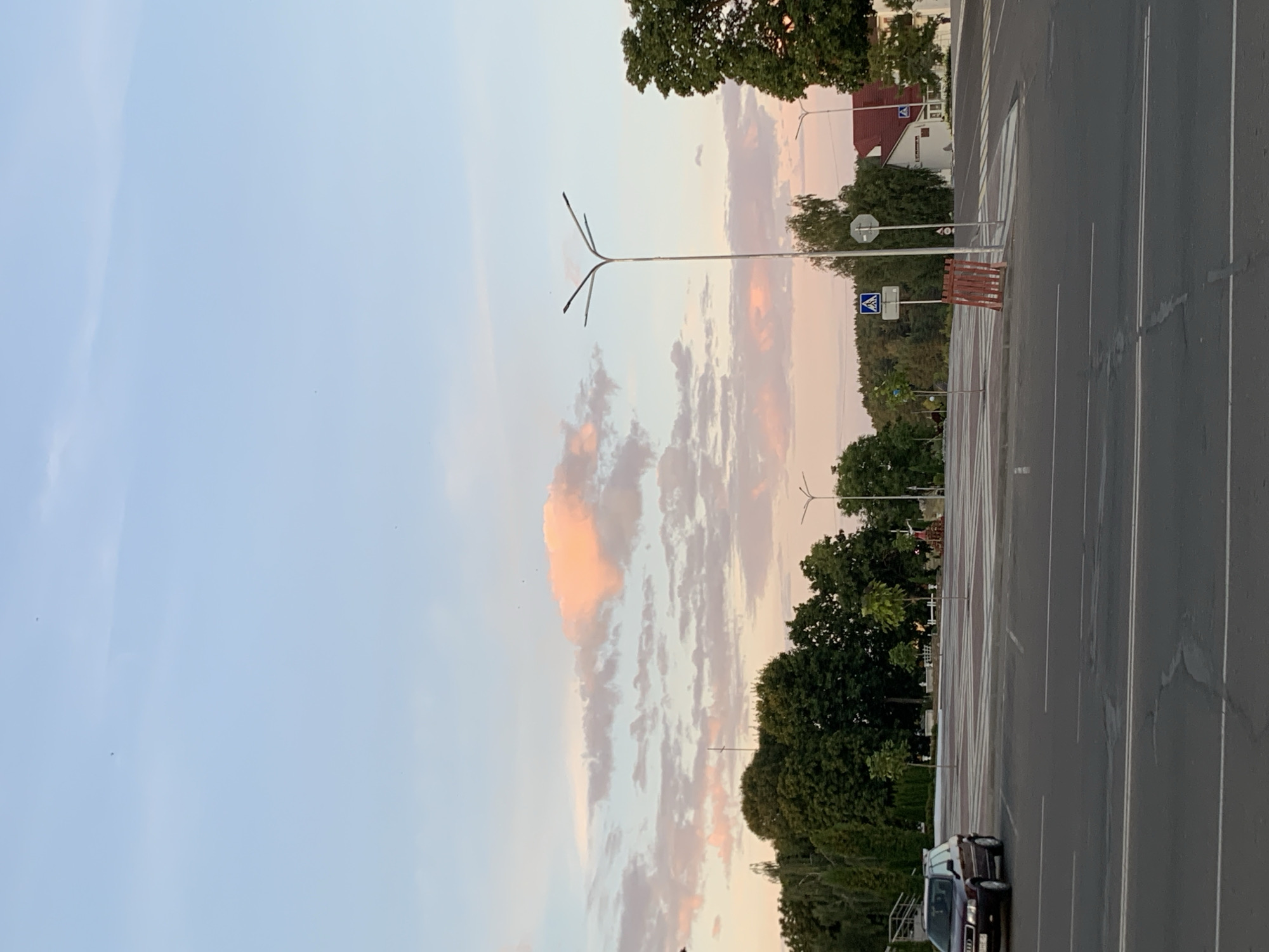 This is a photograph taken in the city of Kapyĺ, Belarus. The image captures a street scene at dusk. The sky is a pale blue with scattered clouds tinged pink and orange from the setting sun. In the foreground, there is an asphalt road with white lane markings. To the left, a dark-colored Audi sedan is parked near a crosswalk. Beyond the road is a large, paved area, possibly a parking lot or plaza, with marked parking spaces. Further back, a line of lush green trees forms a backdrop. A tall, multi-armed lamppost stands in the middle ground. To the right, a building with a red roof is visible, and more trees line the street. Pedestrian crossing signs are present. The overall atmosphere is calm and serene, characteristic of a quiet evening in a city.