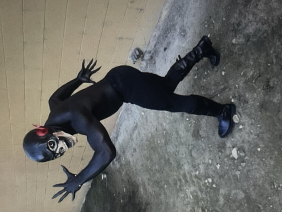 An individual stands outdoors in Muchiya, Nigeria, posed in a dynamic, forward-leaning sprint or lunge. The person is clad in a tight-fitting, all-black costume that covers the entire body. The head is covered by a black mask featuring a white, skull-like face with visible eye sockets, nasal cavity, and teeth, accompanied by red fin-like appendages on either side. The suit includes gloves with elongated, claw-like fingers, with the left arm extended forward and the right arm bent at the elbow. Black, knee-high boots are worn, visible on both feet. The immediate background consists of a light brown or tan masonry wall, likely cinder block, running horizontally. The ground beneath the individual is an uneven, textured surface composed of dark soil, small pebbles, and scattered light-colored debris.
