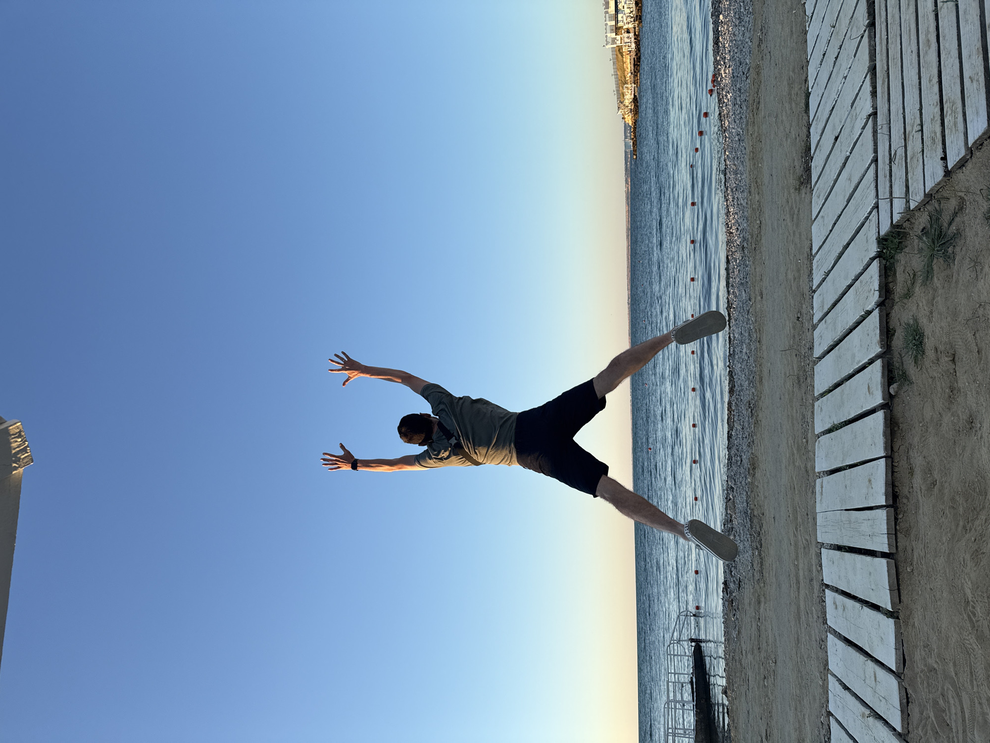 A man is captured mid-jump on a sandy beach with a clear blue sky. He is wearing a dark t-shirt, black shorts, and light-colored sneakers. His arms are raised overhead, and his legs are spread wide, forming an 'X' shape. The ocean stretches out behind him, with a distant shoreline featuring buildings and structures visible on the right. In the foreground, a weathered wooden boardwalk is laid out on the sand. The lighting suggests it is either early morning or late afternoon, with the sun casting a warm glow.