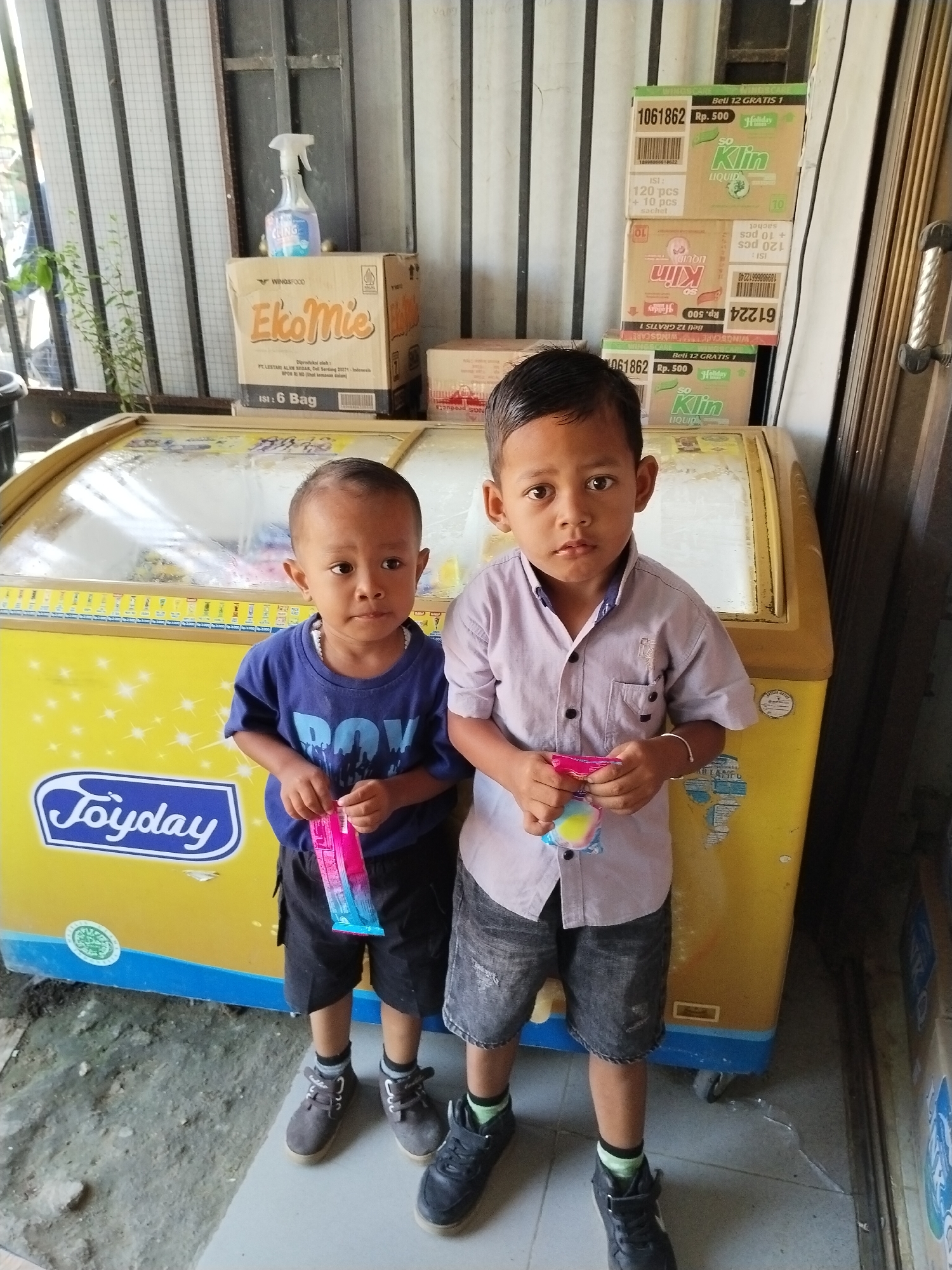 Two young boys stand in front of a yellow and blue commercial freezer displaying a "Joyday" logo. The boy on the left wears a dark blue t-shirt and black shorts, holding a pink and blue popsicle. The boy on the right wears a light purple collared shirt and dark grey denim shorts, also holding a colorful popsicle. Both boys are wearing sneakers. Behind them, stacked boxes are visible, including "EkoMie" noodles and "So Klin" liquid detergent, along with a spray bottle of cleaner. The background appears to be an indoor/outdoor space with a metal grate partition and vertical wooden planks. The overall scene suggests a retail environment in Sawit Seberang, Indonesia.