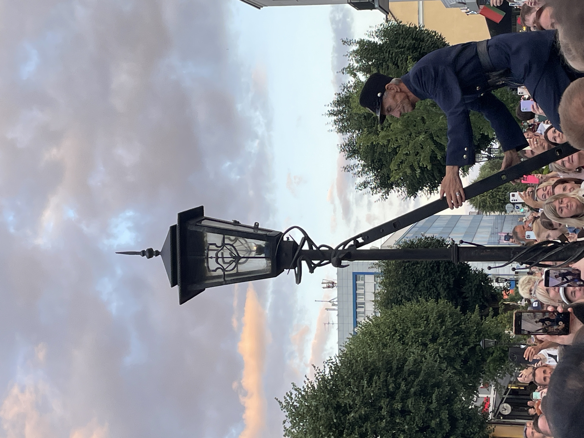 This media file captures a street scene in Brest, Belarus, likely during an outdoor event. The main subject is a man dressed in a historical-style dark blue uniform with a cap, leaning over a black lamppost. He appears to be a performer or historical reenactor, given his attire and the audience gathered around.
The scene is populated by a crowd of people, many of whom are holding up smartphones, suggesting they are documenting the event. The time of day appears to be late afternoon or early evening, as indicated by the soft, diffused light and the pastel colors in the cloudy sky. The weather seems mild and overcast, with some areas of the sky showing soft pink and orange hues from the setting sun.
The lamppost itself is an ornate, dark metal structure with a glass lantern, a common feature in older European city streets. Behind the lamppost and the performer, lush green trees are visible, along with a modern building with a glass facade. The presence of trees and the style of the architecture suggest an urban park or a public square.
The man's facial expression, with his furrowed brow and focused gaze, conveys concentration as he interacts with the lamppost. The people in the crowd appear engaged and curious, with some smiling as they take photos. The overall atmosphere seems festive and engaging, characteristic of a public performance or festival. No visible text is clearly discernible in the image.