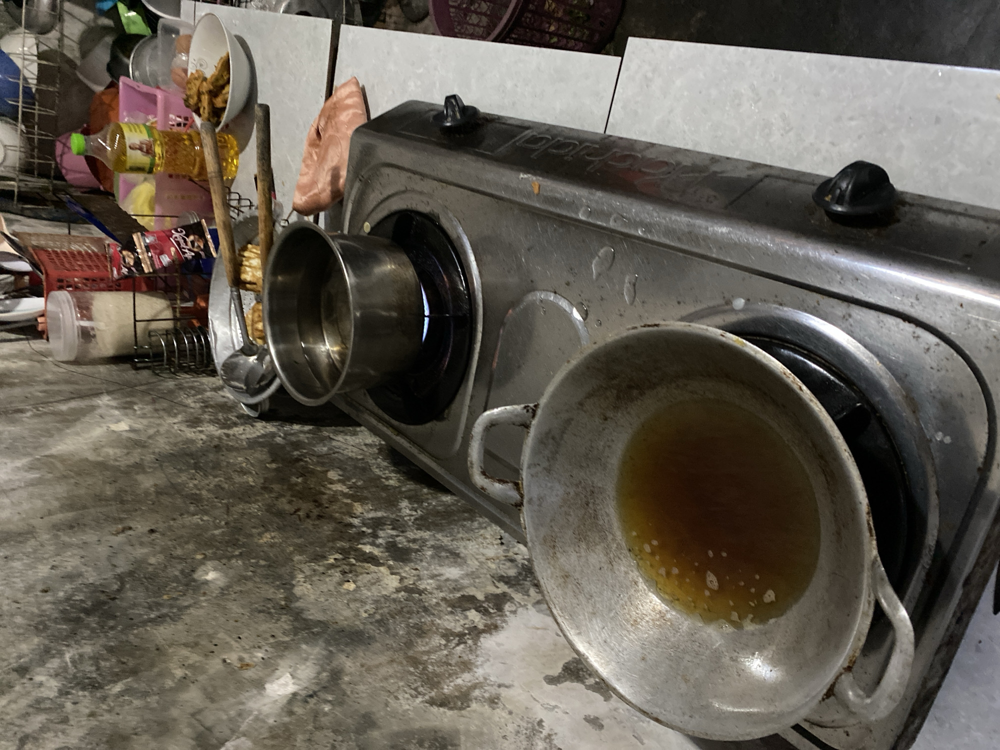 A still life composition captures a cluttered kitchen scene, likely within the city of Madiun, Indonesia. The foreground is dominated by a well-used aluminum wok filled with golden-brown cooking oil, placed on the left burner of a silver gas stove. The right burner of the stove holds a small stainless steel pot containing clear liquid, with a blue flame visible beneath it, indicating it is actively heating. To the right of the stove, a white bowl contains several pieces of fried food. Behind the stove, a bottle of cooking oil stands prominently. The background reveals a textured, somewhat grimy concrete wall. Various kitchen items, including utensils in a red basket and containers, are visible on a tiled countertop, adding to the lived-in atmosphere of the space.