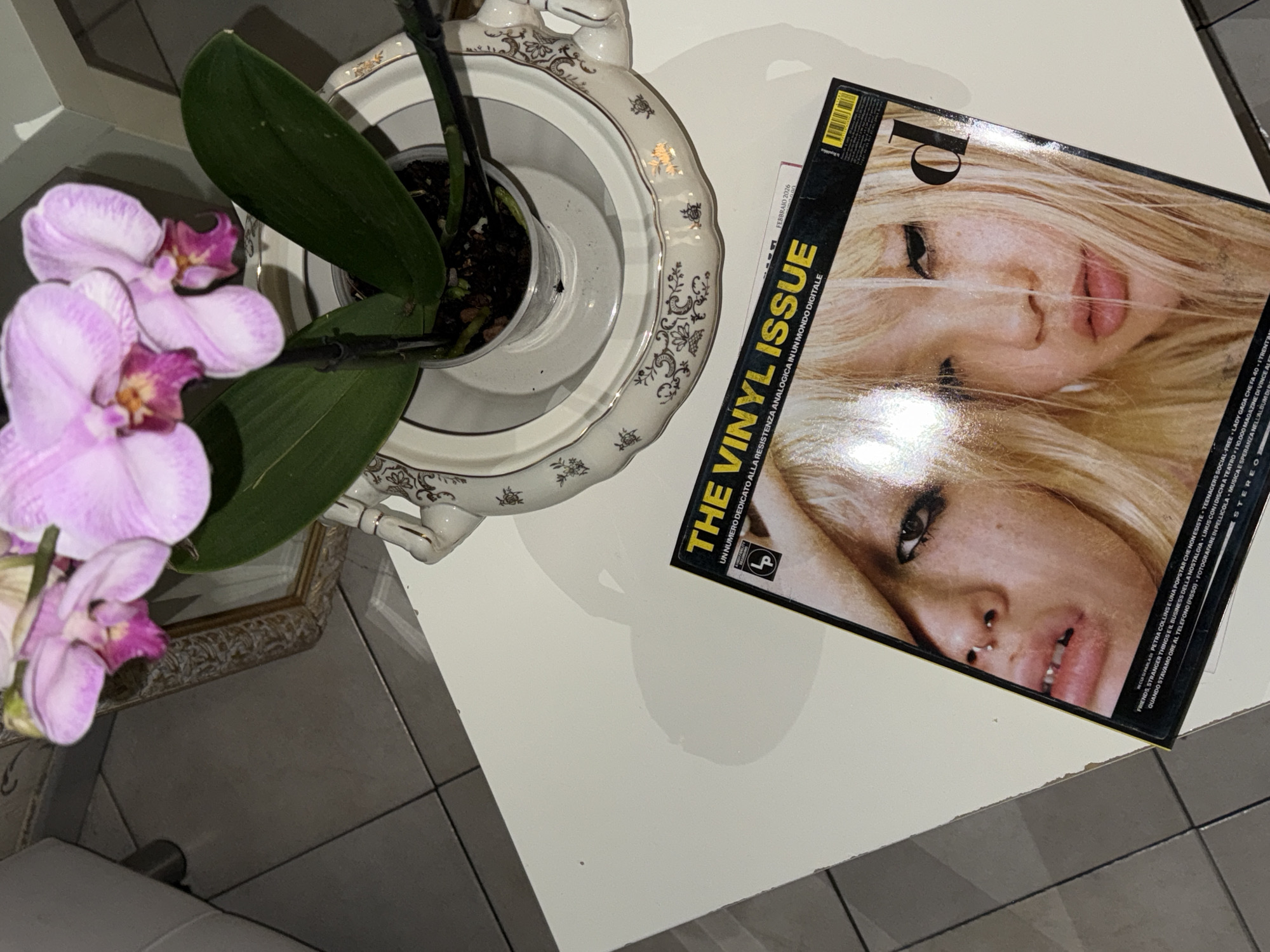 The image depicts a top-down view of a table setting, likely indoors, potentially in Sanremo, Italy given the Italian text on the magazine. On the left is a light pink orchid plant in a pot sitting inside an ornate, decorative white bowl, possibly porcelain. To the right of the plant, on the table, is a copy of "The Vinyl Issue" of a magazine, with a blurred image of two blonde women on the cover. The table is a white, flat surface. The surrounding environment includes tile flooring visible in the lower-left and lower-right corners. A partially visible picture frame can be seen on the lower-left side of the frame. The scene is well-lit. There are no people directly visible within the frame.