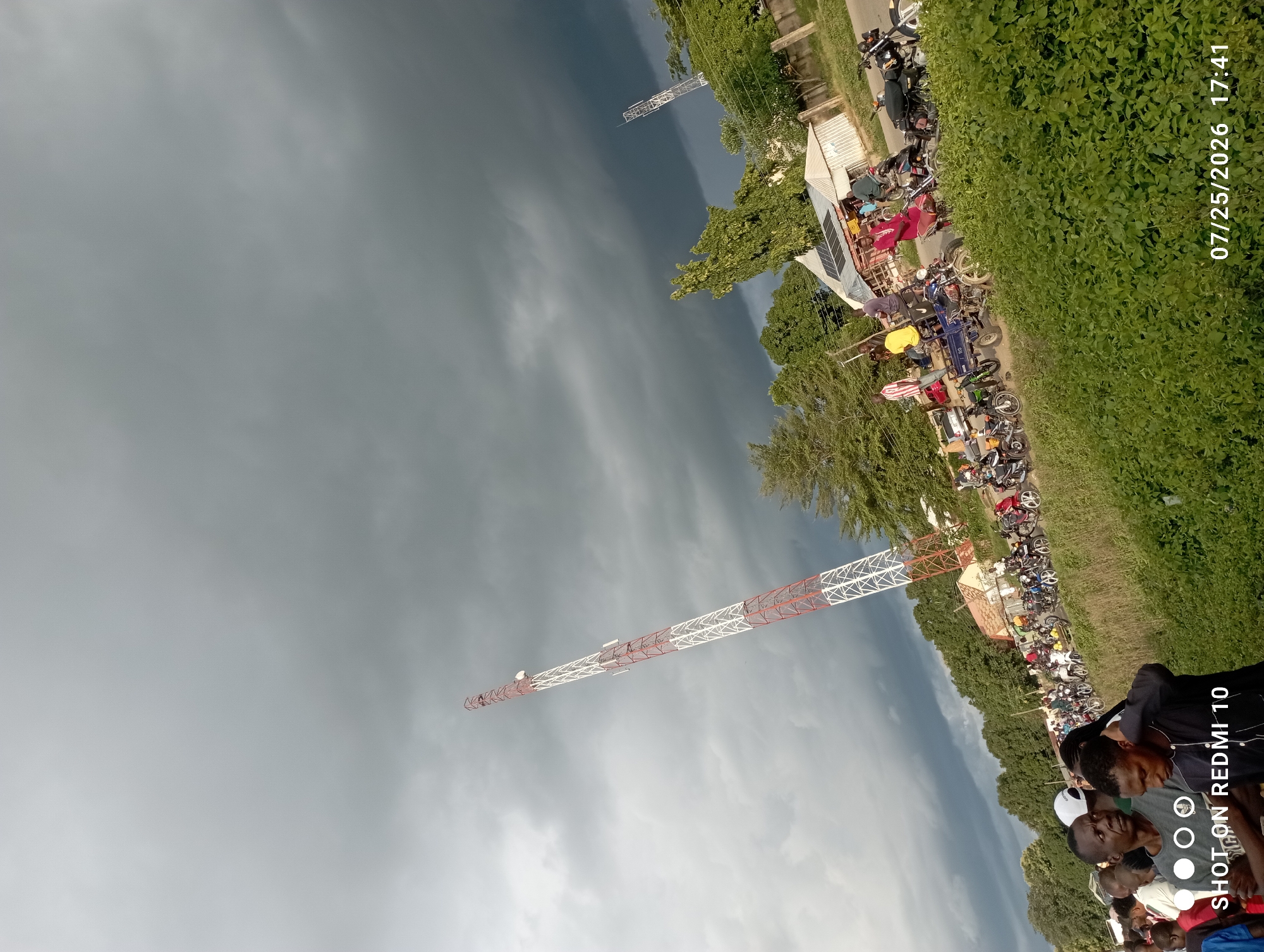In Serti, Nigeria, an outdoor gathering is underway under a stormy, dark gray sky. A tall, red and white telecommunications tower dominates the left side of the frame, angled upwards. A line of people, mostly men, are gathered on the left side of a dirt road, facing towards the right. On the right side of the road, numerous motorcycles are parked in rows, with several individuals standing or sitting near them. Some of these individuals appear to be interacting. A building with a corrugated metal roof and solar panels is visible in the middle ground, situated behind the motorcycles. Trees and vegetation surround the area, with lush green bushes in the foreground. The image is dated 07/25/2026 at 17:41.