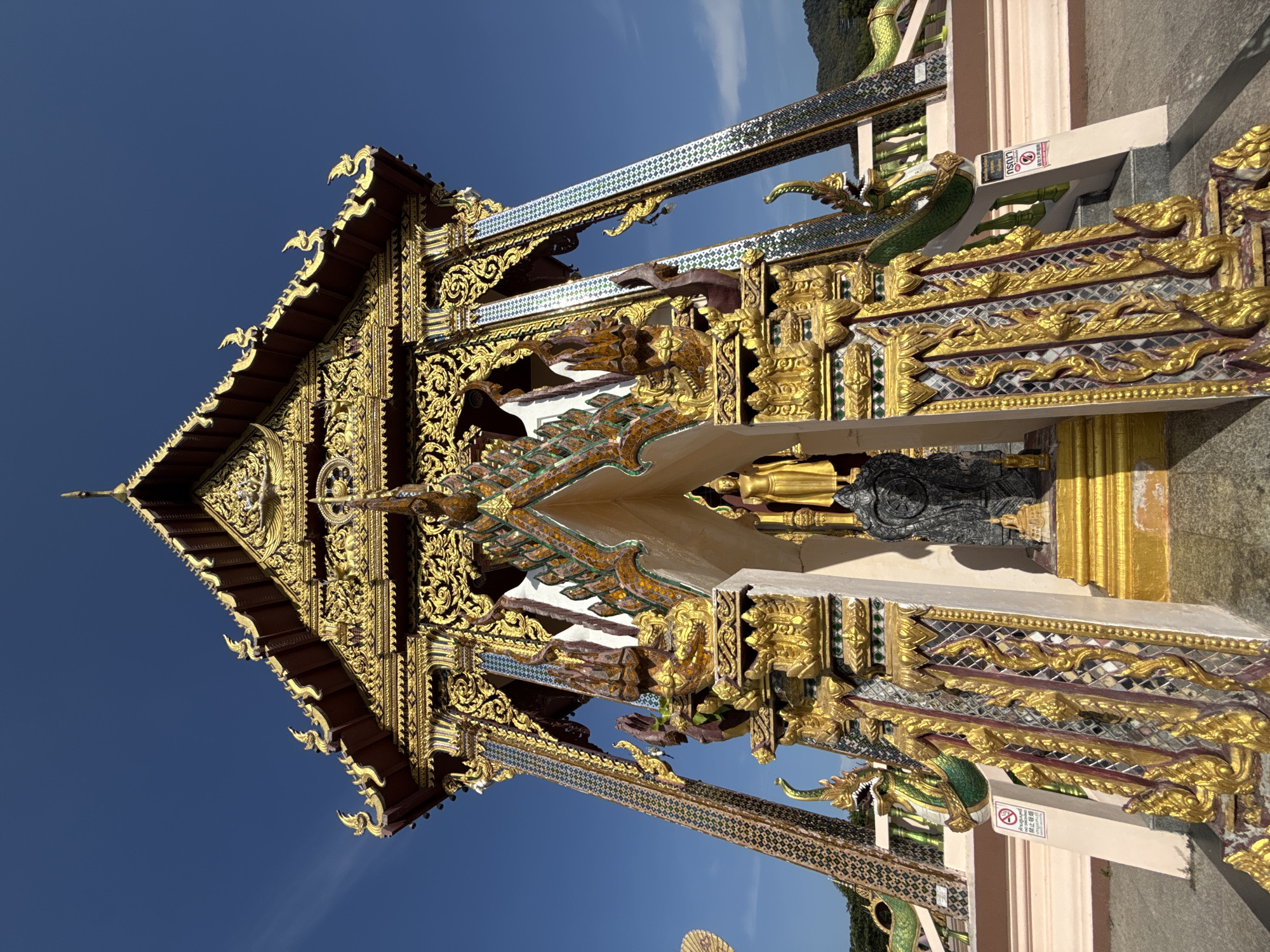 This image shows a low-angle, exterior shot of a highly ornate Buddhist temple, likely in the city of Ko Samui District, Thailand. The temple features elaborate golden carvings, intricate mosaic tile work in shades of blue, green, and gold, and is topped with a multi-tiered, pointed roof adorned with decorative elements. A large, golden Buddha statue stands prominently within the temple's entrance, partially obscured by a dark, possibly stone, statue in front of it. Flanking the entrance are ornate pillars and decorative serpent-like statues. The scene is set against a clear, bright blue sky, with hints of green foliage and distant hills visible in the background. The ground in the foreground appears to be paved.