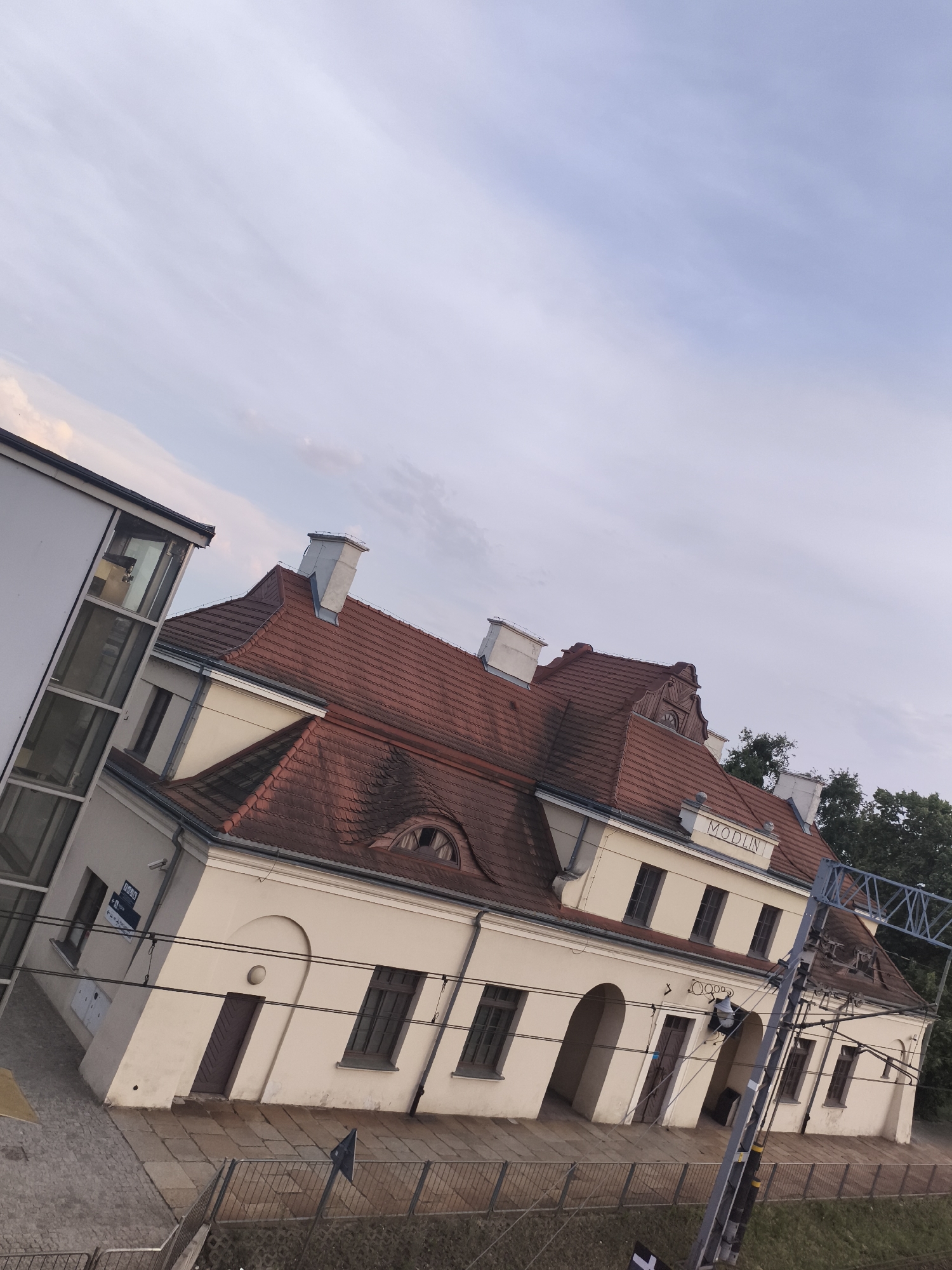 The image shows a light-colored building with a red tile roof, likely a train station, with the name "MODLIN" visible in raised lettering on the facade. A modern glass-enclosed structure is attached to the left side of the building. The scene is angled, suggesting it was taken from an elevated position or the camera was tilted.
The building has multiple windows, some arched, and several chimneys. There are overhead electrical wires and a metal support structure in the foreground, typical of a railway environment. The sky is mostly cloudy with a pale, diffused light, indicating it is likely late afternoon or early evening. Some trees are visible behind the building on the right. The ground in front of the building appears to be paved with stone slabs and cobblestones. There is a metal fence along the platform.
A sign with Polish text and symbols is visible on the left side of the building. The overall impression is one of a somewhat old, yet functional, public building in a semi-urban or suburban setting. No people are visible in the image. The lighting and the cloudy sky contribute to a serene, calm mood.