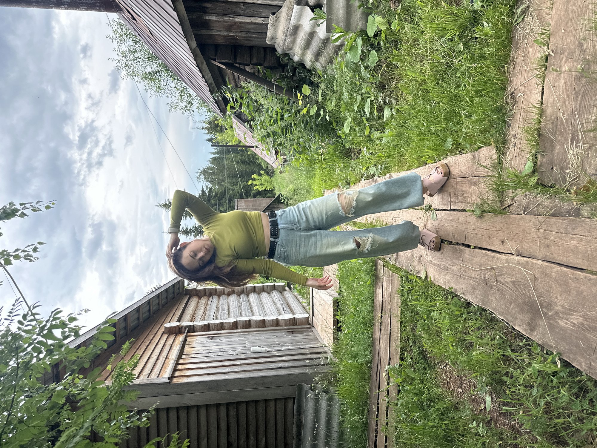 A young woman with long brown hair is lying down on a wooden path in what appears to be a rural setting. She is wearing a light green long-sleeved crop top, distressed light blue jeans, and light brown sandals. The woman is looking upwards with her eyes closed and her head resting on her hand, suggesting a moment of relaxation or rest. Her pose is casual and somewhat sprawled.
The surrounding scene is lush and green, with tall grass and various plants growing. Behind her, there is a rustic wooden building, possibly a shed or a small house, with weathered wooden planks. Another building, also wooden and with a corrugated roof, is partially visible on the left. The path is made of rough-hewn wooden planks, laid out in a somewhat uneven manner. The sky above is partly cloudy, with a mix of white clouds and blue sky visible. The overall impression is one of a peaceful, outdoor moment in a natural, possibly slightly unkempt, environment. The time of day appears to be daytime, likely late morning or afternoon, given the lighting. No visible text is present in the image.