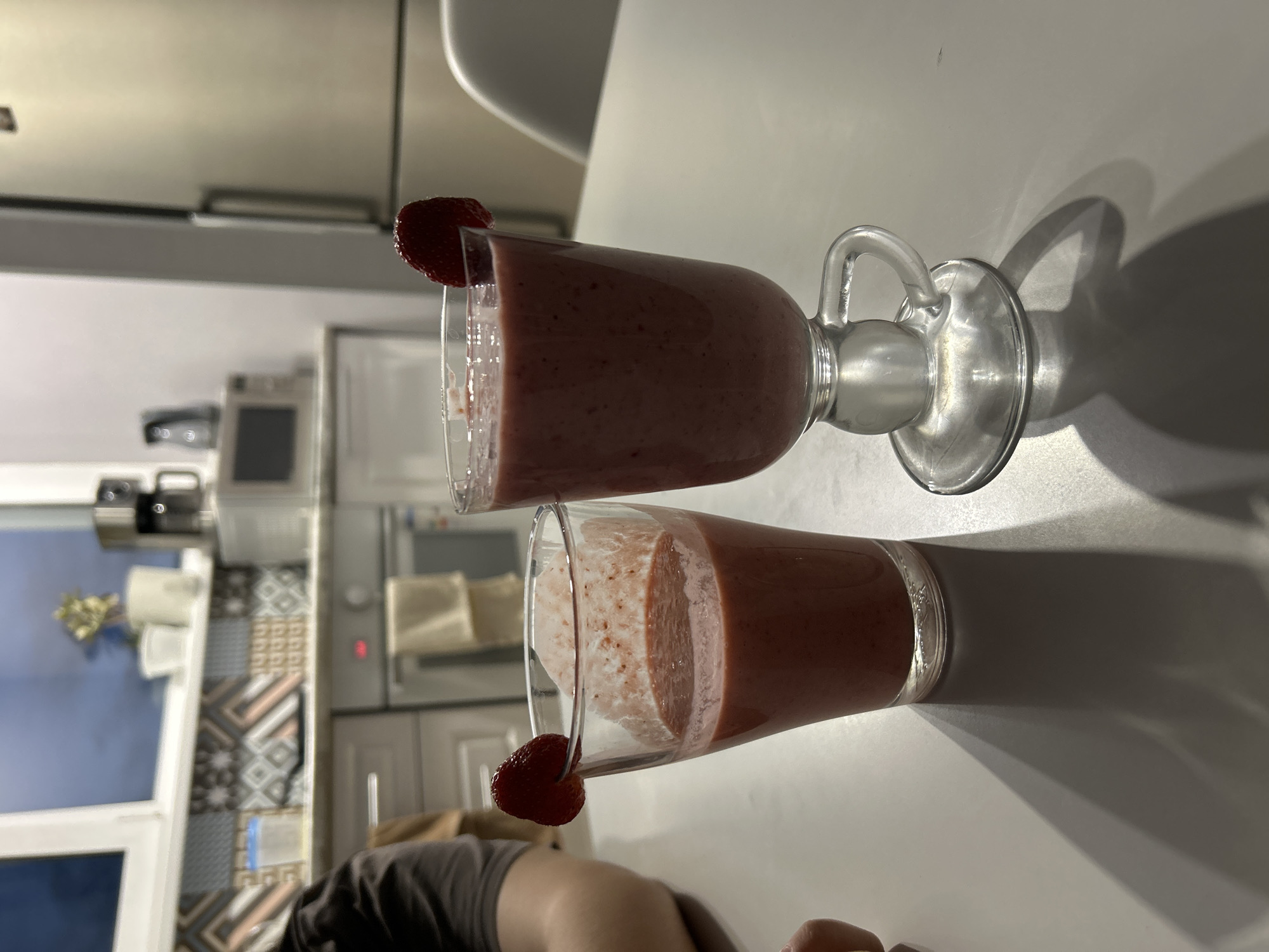 Two glasses of strawberry smoothie sit on a white table in what appears to be a kitchen. Each glass is garnished with a strawberry slice on the rim. The smoothie is a pale pink color with visible flecks of strawberry seeds. One glass is a stemmed glass with a handle, while the other is a more traditional tumbler. The background shows kitchen cabinets, a tiled backsplash, and a window. The lighting suggests it could be daytime, with shadows cast by the glasses. The overall scene is casual and domestic, possibly implying a refreshment being enjoyed at home. No visible text is present in the image.
