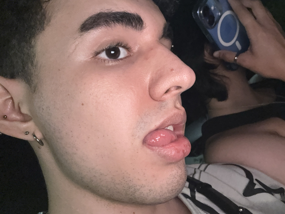 A close-up shot reveals a young man with dark hair and eyebrows, brown eyes, and fair skin. His tongue is extended, slightly protruding from his lips. He has multiple piercings on his ear. A blurred figure behind him holds a blue cell phone, partially obscuring the background. The man wears a white and black striped shirt. The image showcases the man's facial features and expression in detail.