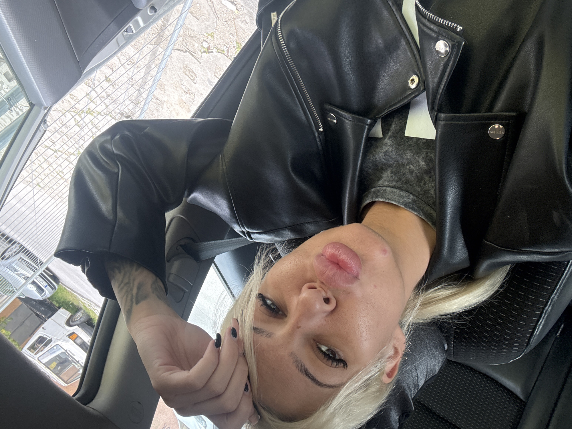 A blonde woman in a black leather jacket is in a car, holding her hand near her face. She's looking at the camera. The setting appears to be in a city, judging by the vehicles visible through the car window.