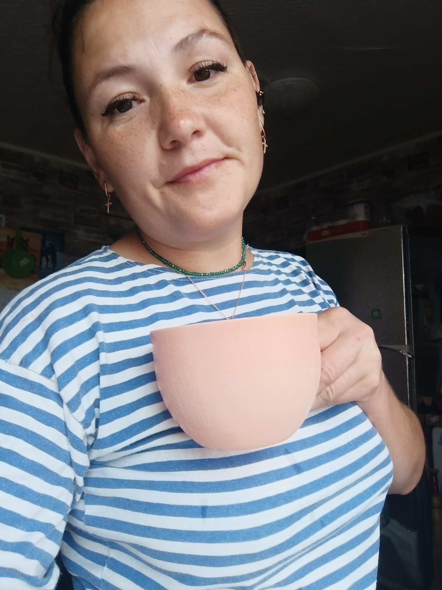 A woman with freckles and dark hair is holding a pale pink cup. She is wearing a blue and white striped shirt and a green beaded necklace. Her right hand is holding the cup, and her left hand is not visible. She has earrings shaped like crosses. The background is dark and appears to be a kitchen, with a refrigerator visible on the right. The image is a selfie, taken from a slightly low angle. The location is not specified as Karsha, Russia.